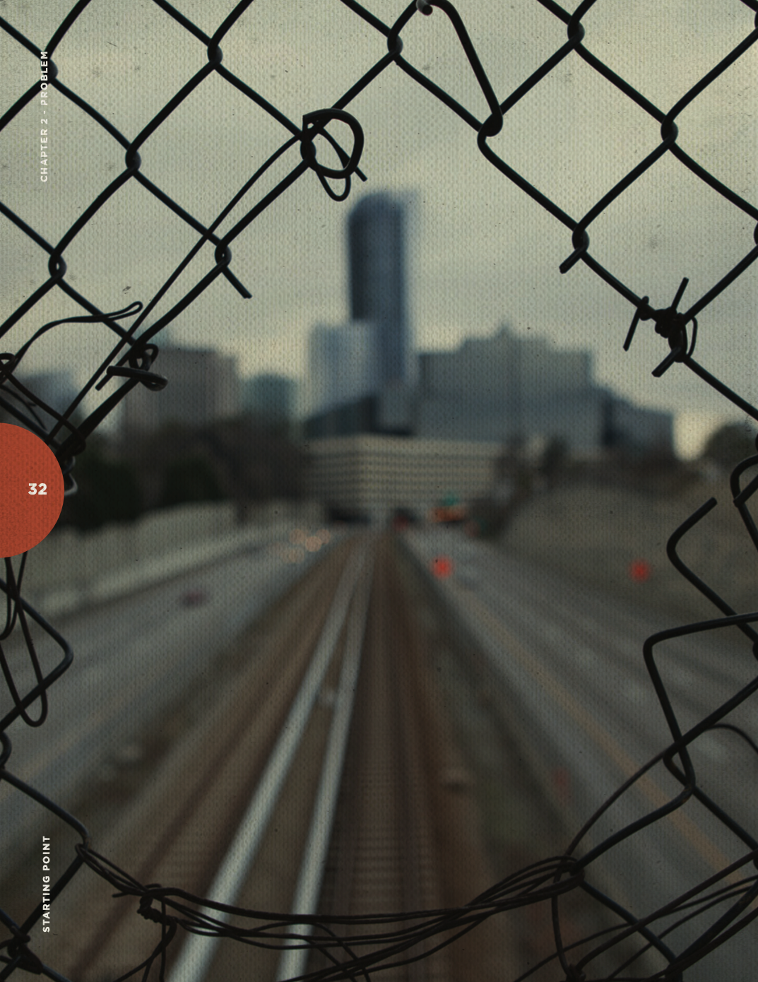 A view of a city skyline through a damaged chain-link fence with a railway running along the foreground.