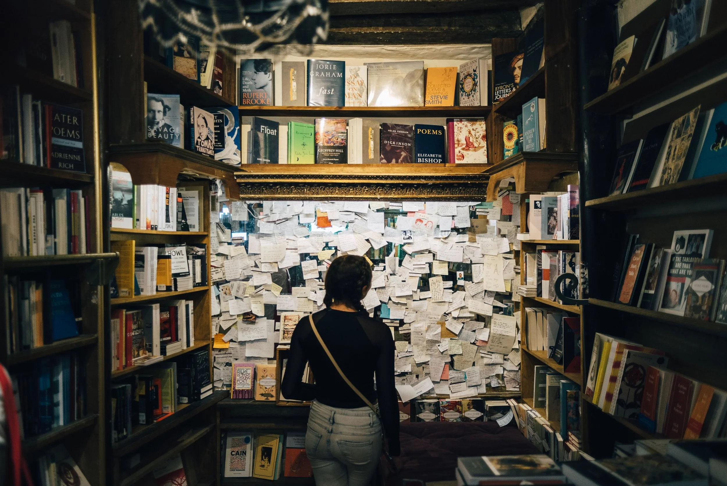 A woman with dark hair, wearing a black long-sleeve top and light-colored jeans, stands in a bookstore reading area, facing a wall covered with numerous notes and papers. Bookshelves filled with books surround her. The top shelf displays books with t