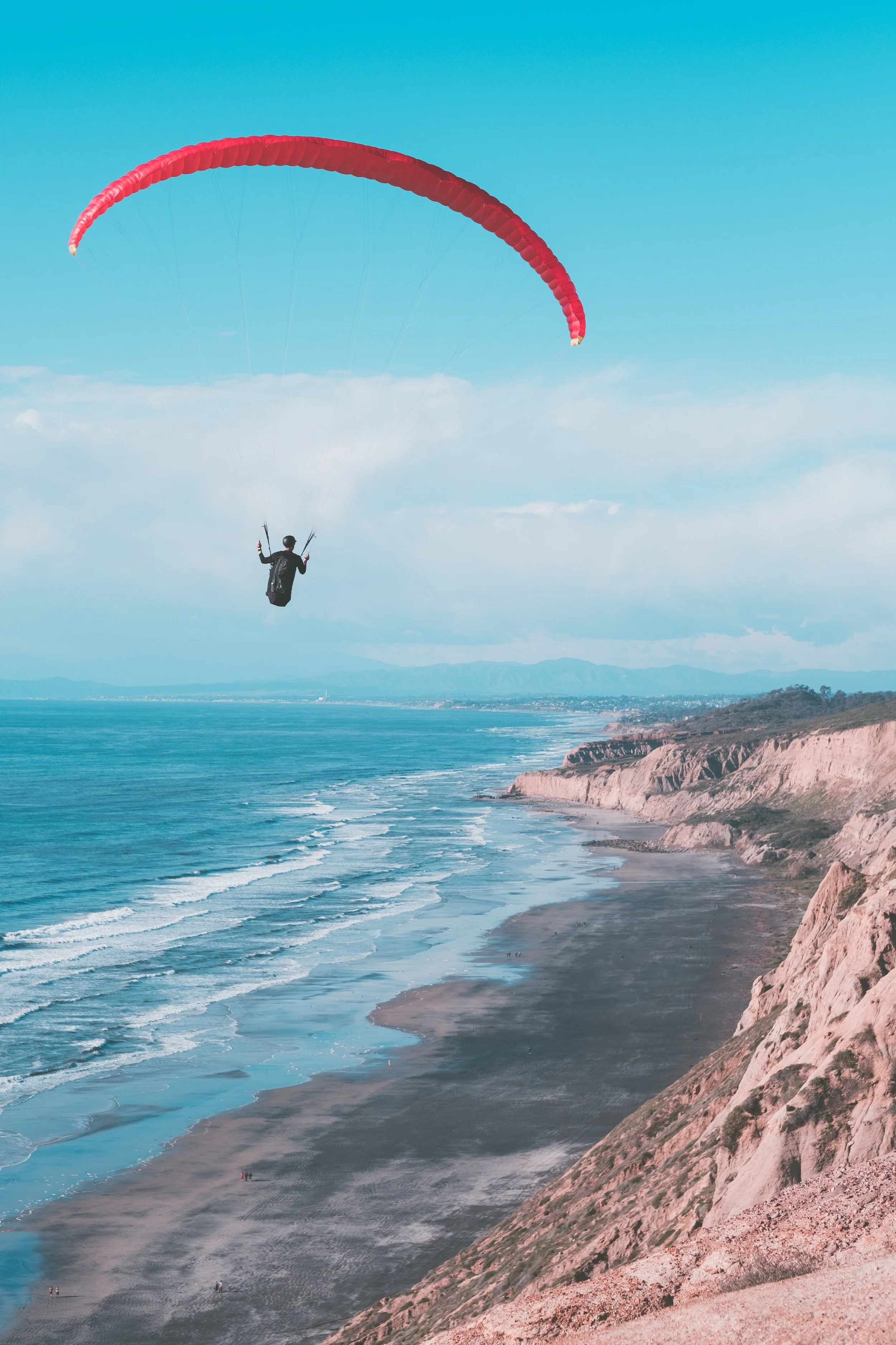 A person paragliding over a beach with cliffs, ocean waves, and a clear sky.