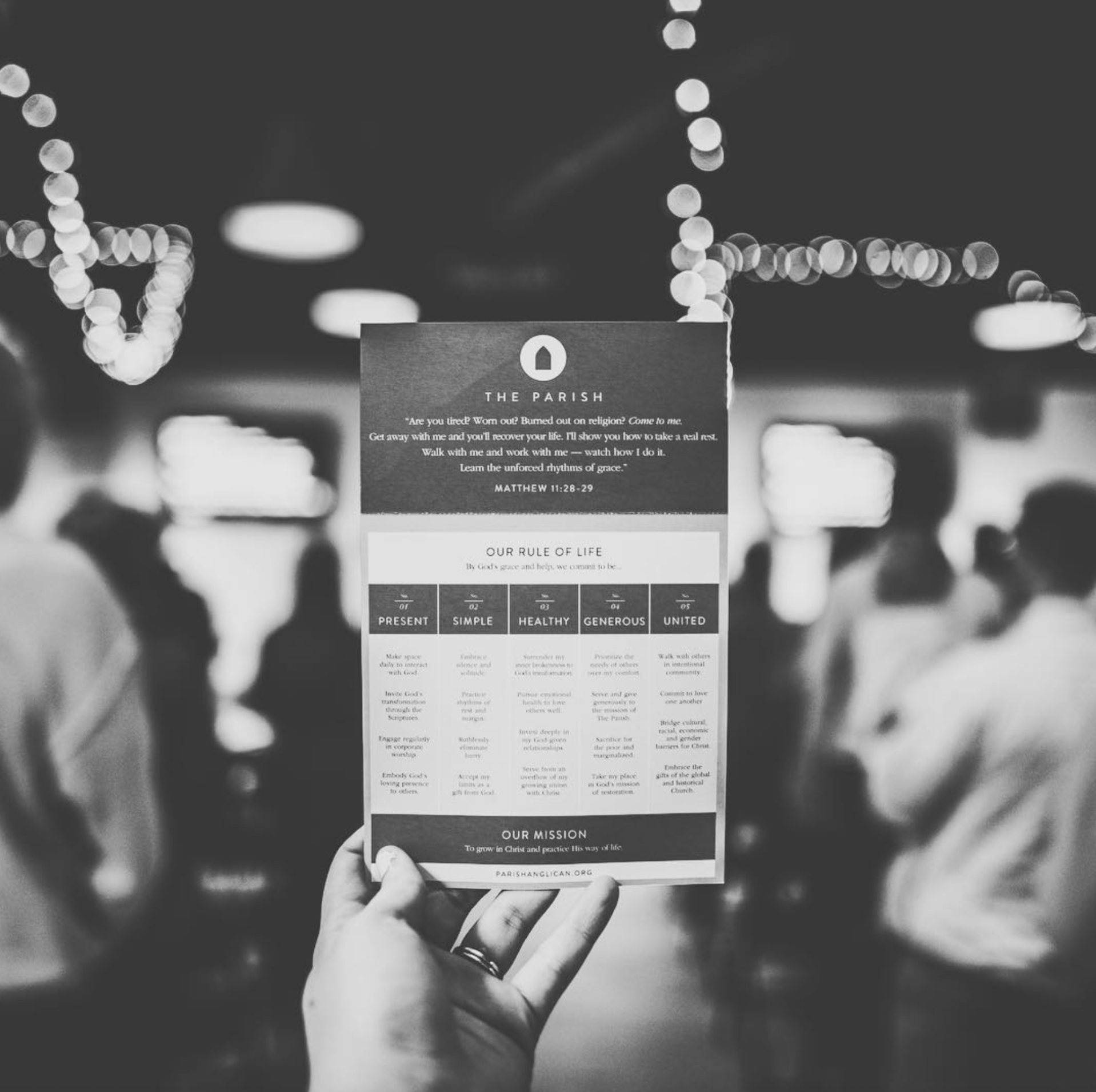 A hand holding a flyer in a church setting with people seated in the background. The flyer contains religious text and rules of life for a parish.