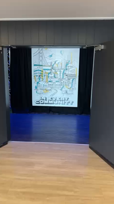 View of a small stage with a screen displaying a cityscape illustration and the words "In Every Community" at the bottom.