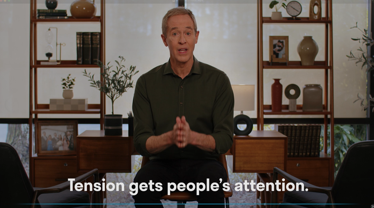 A man with gray hair in a green shirt sitting on a wooden chair, speaking in a room with shelves, framed pictures, vases, and a lamp behind him. A caption at the bottom reads, "Tension gets people's attention."