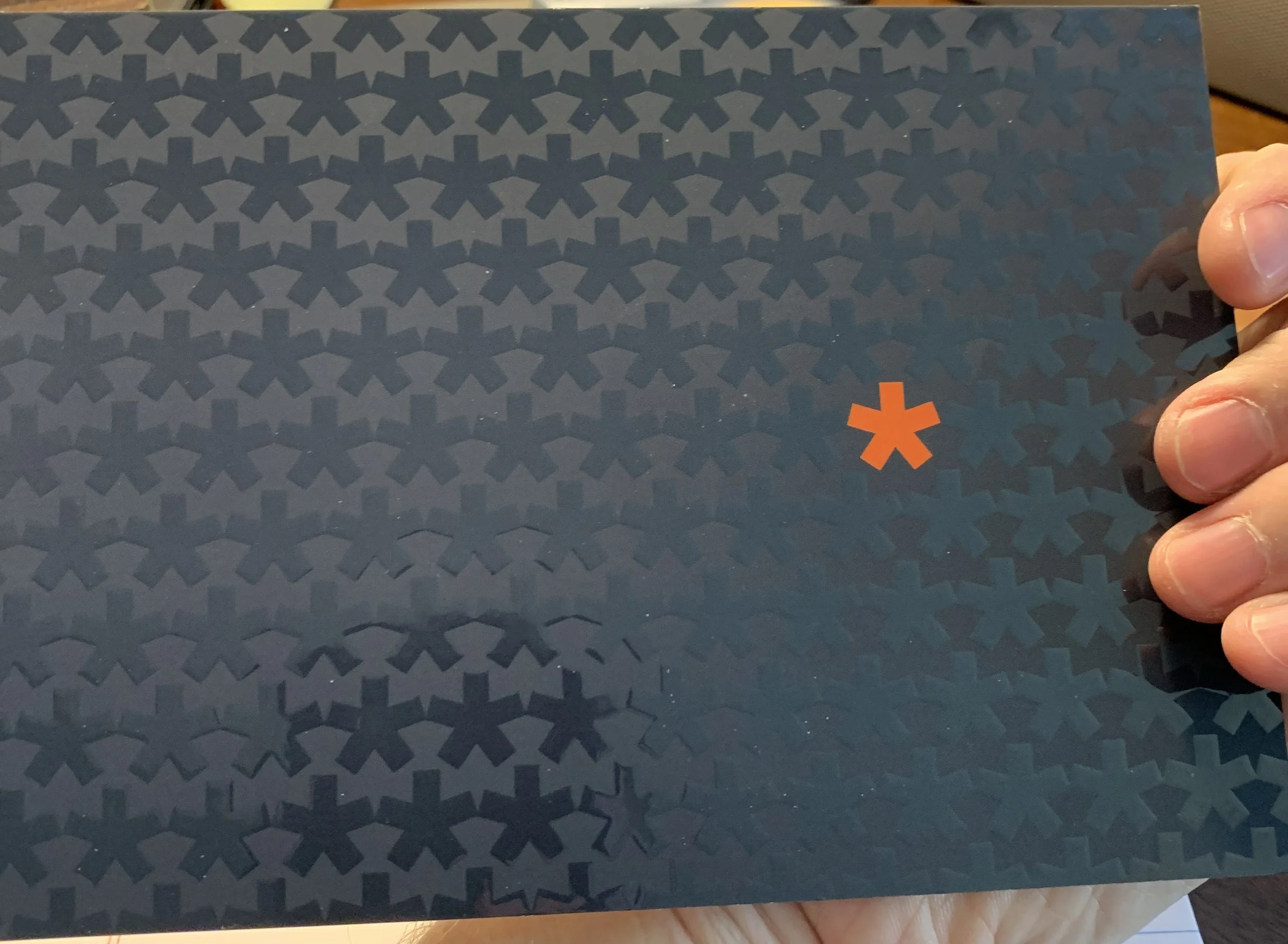 A dark background with a repeating pattern of X shapes and a small orange asterisk symbol on the right side, held by a person's hand.