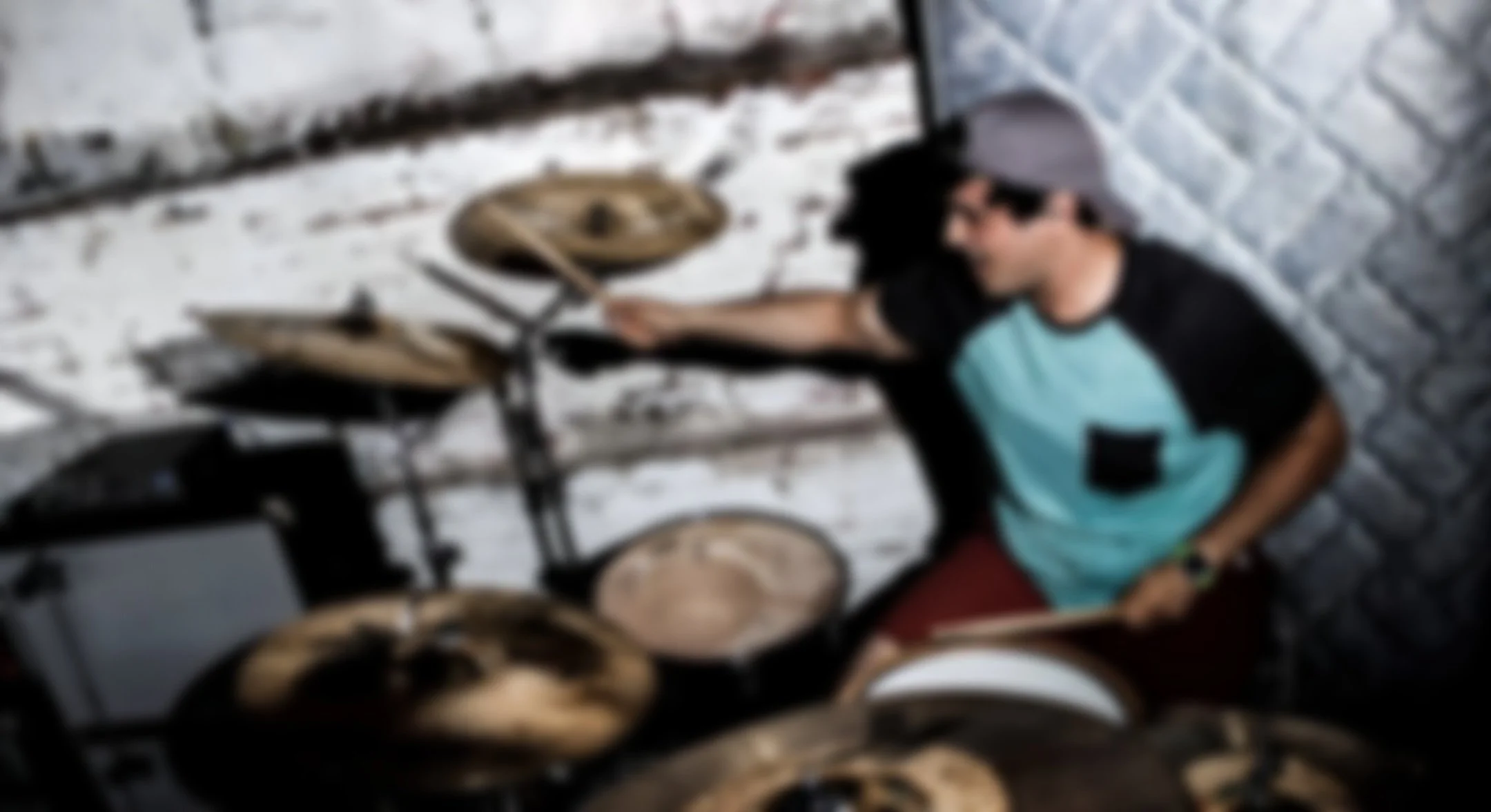 A young man wearing a baseball cap, t-shirt, and watch is playing a drum set in an industrial-style room with a white brick wall and a padded silver wall panel.
