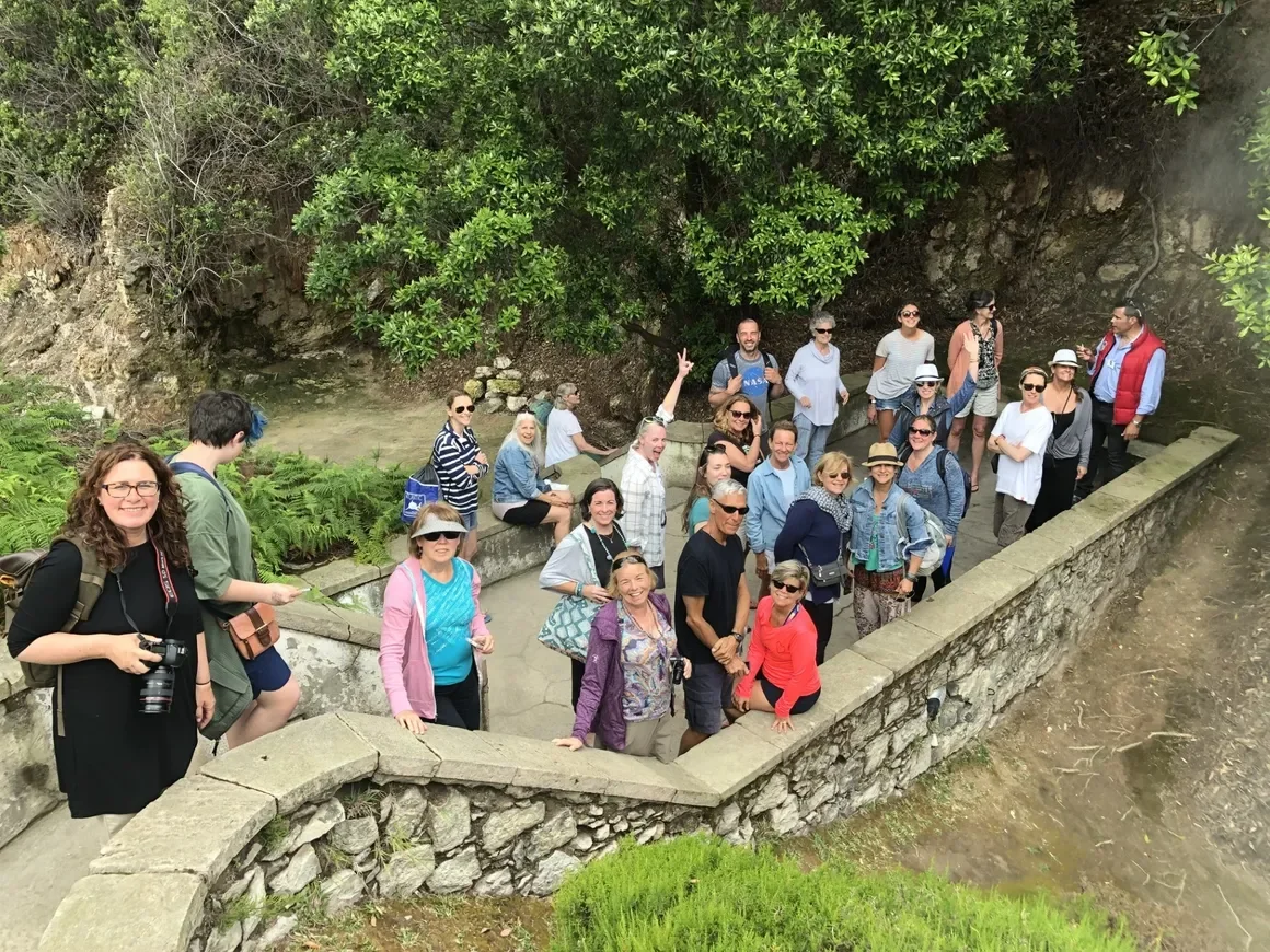 Group of people gathered on a stone pathway with lush green trees and bushes behind them, some sitting on the stone wall, others standing and smiling, in a the outdoor setting of Azores, Portugal. Hiking in the Azores with a group of yoga students.