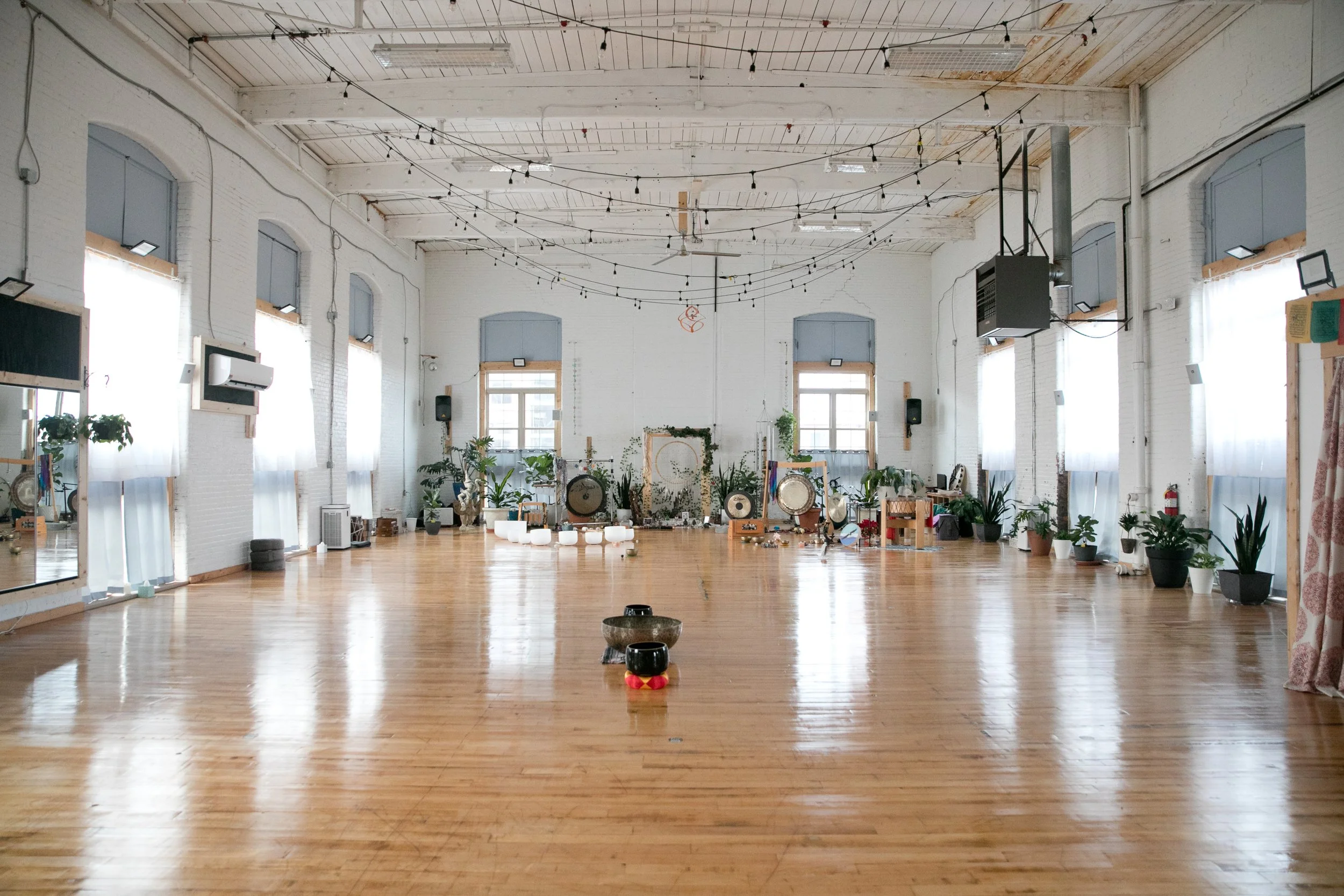 An empty, spacious dance or event studio with wooden floors, white brick walls, large windows with sheer curtains, and string lights hanging from the ceiling. The back wall features plants, drums. There are mirrors on the left wall and various small objects scattered around.