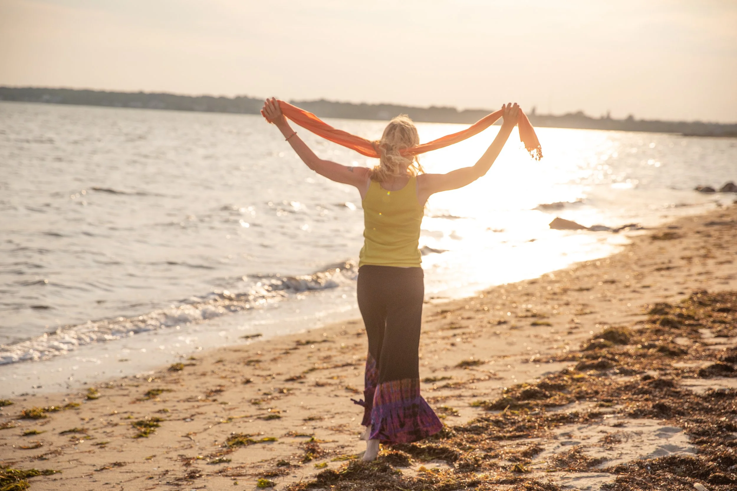 A woman standing on a beach holding an orange scarf over her head during sunset.