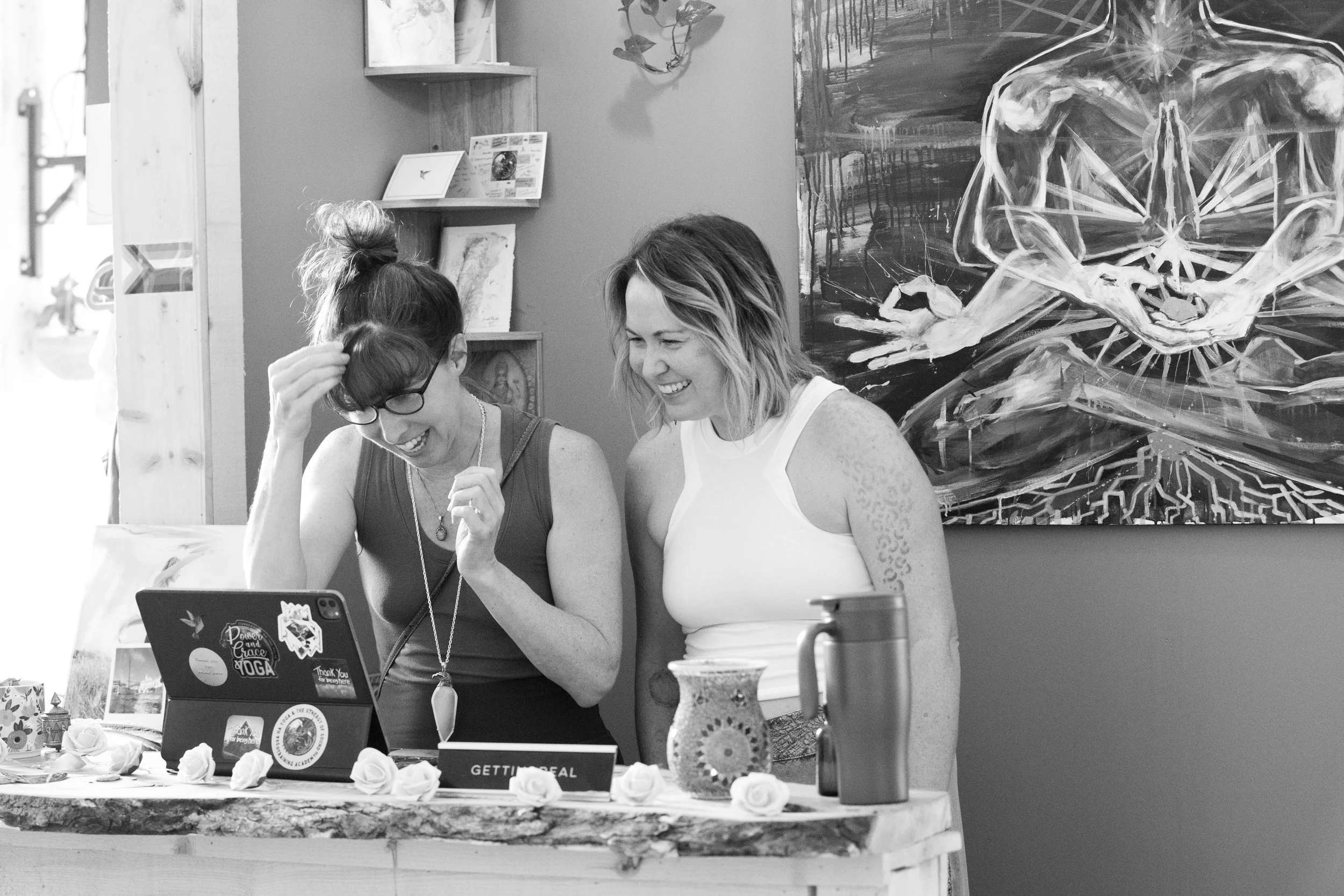 Two women laughing and looking at a laptop at a table with decorative items, flowers, and a sign that says "Getting Real". Art painting and shelves are visible on the wall behind them.