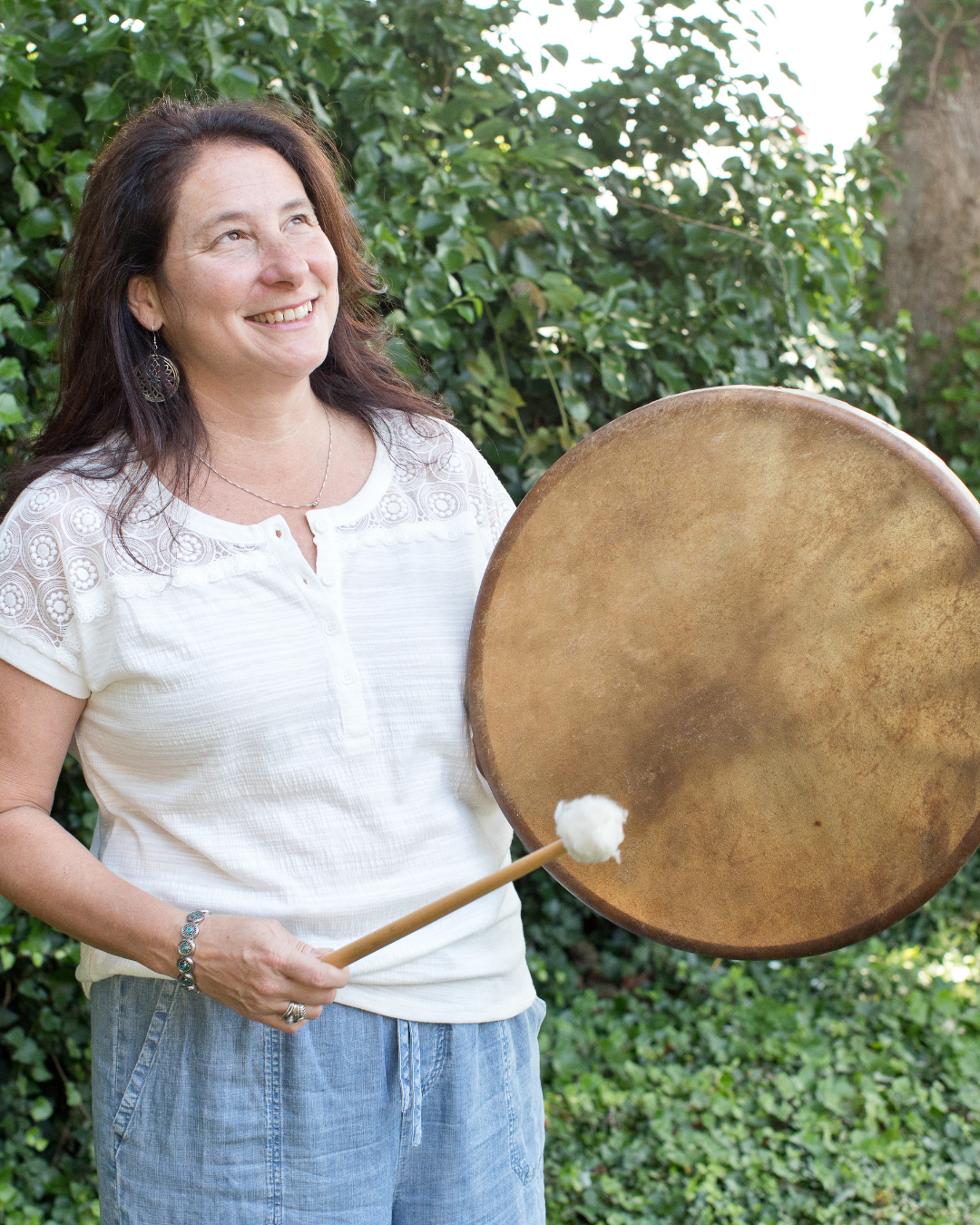 A woman smiling outdoors, holding a large Tibetan singing bowl with a mallet, standing in front of greenery.