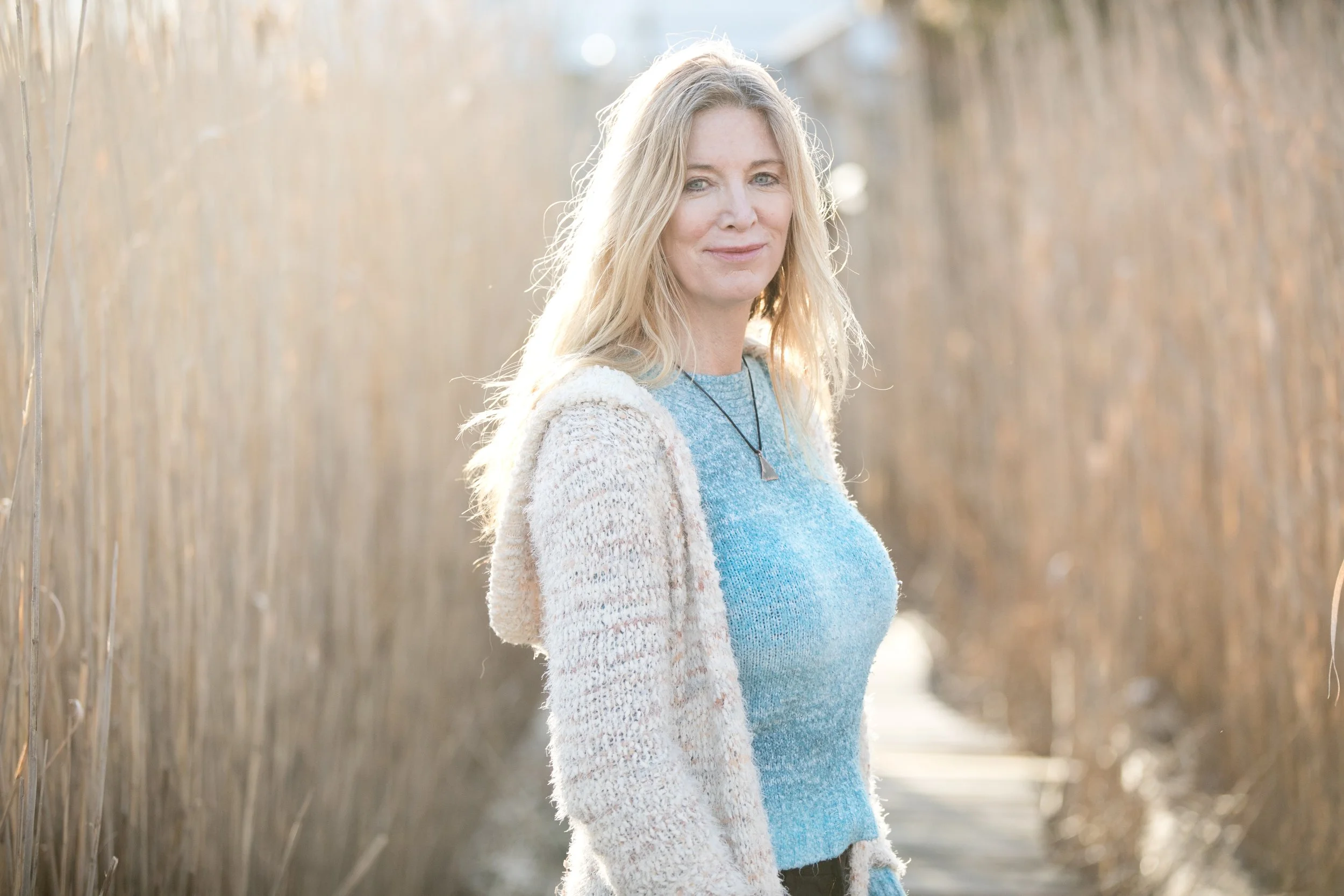 A woman with long blonde hair standing on a narrow wooden pathway surrounded by tall dry grass, with sunlight illuminating her face and hair.