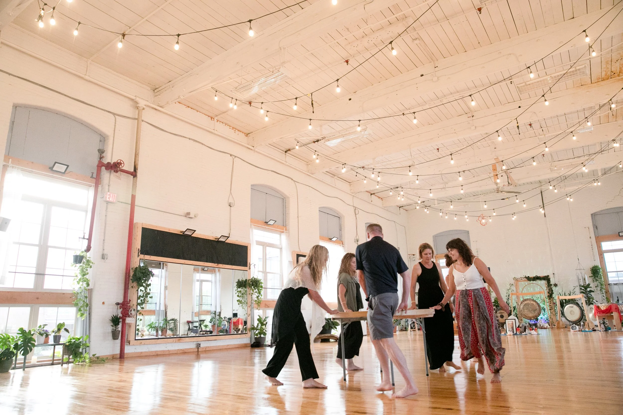 Group of people in Sva Ha Yoga studio channeling with the OHM Drum, with string lights hanging from the ceiling and potted plants near the windows.