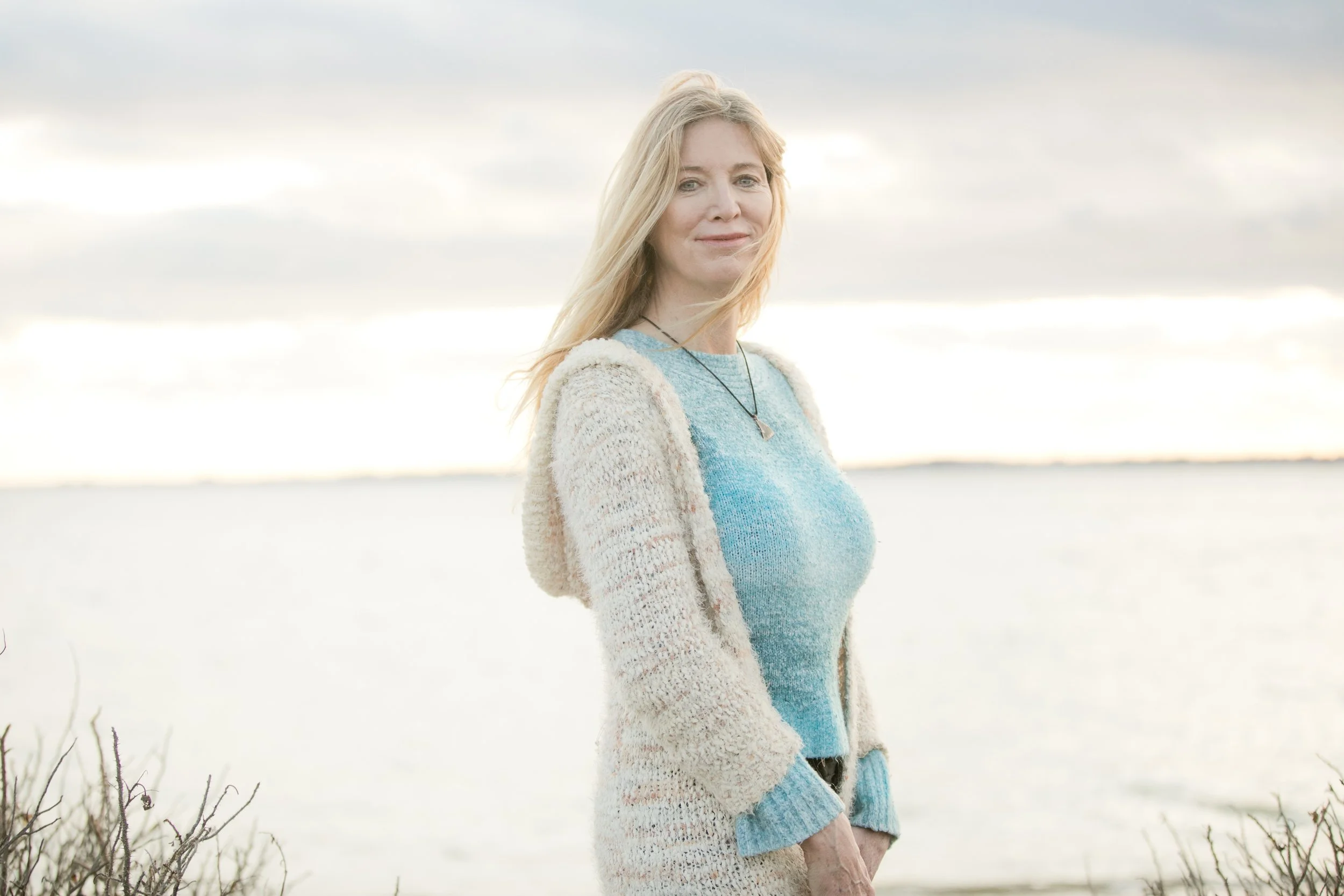 A woman with blonde hair standing outdoors near a body of water during cloudy weather.