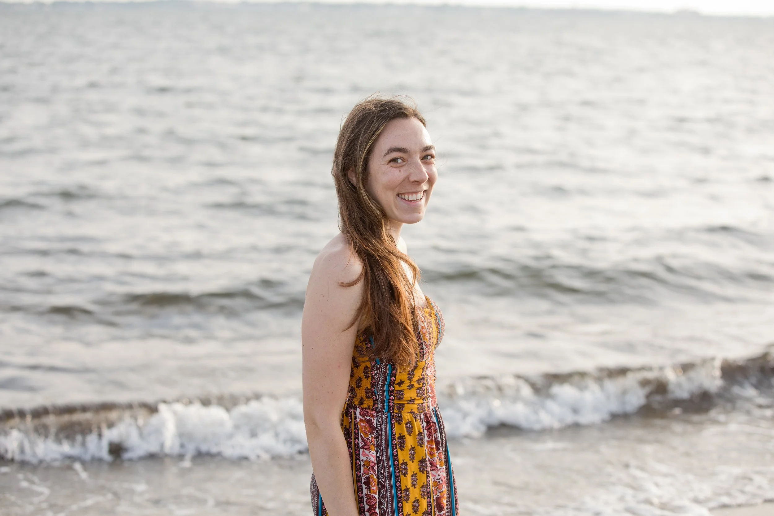A young woman with long brown hair standing on a beach with water and small waves in the background, smiling at the camera.