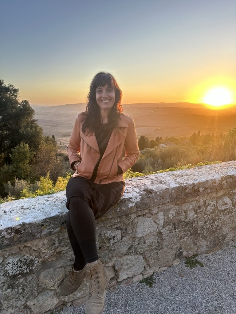 A young woman with dark wavy hair, smiling, sitting on a stone wall during a sunset in the countryside with rolling hills and trees in the background.