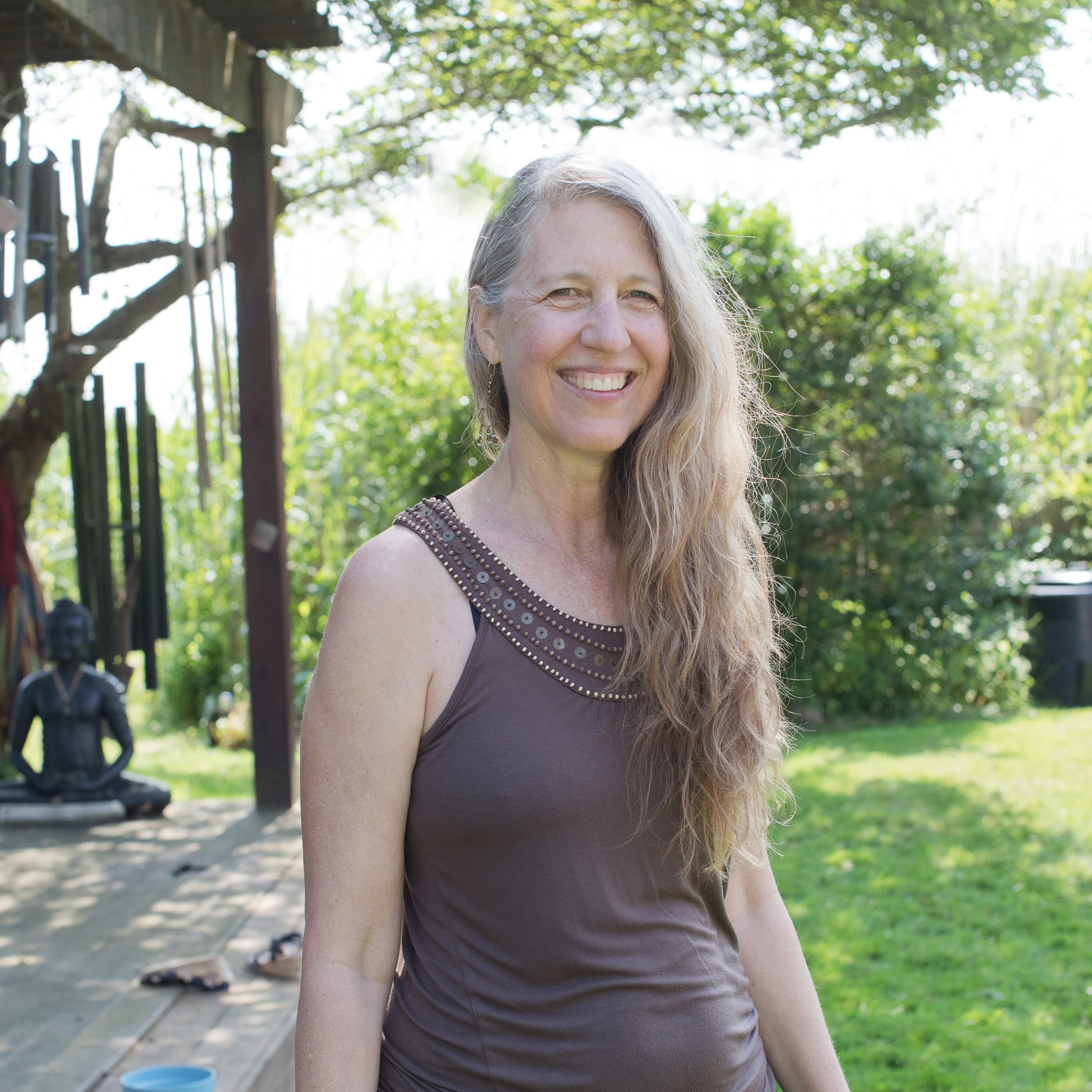 Smiling woman with long, wavy hair standing outdoors on a sunny day, with trees and greenery in the background.