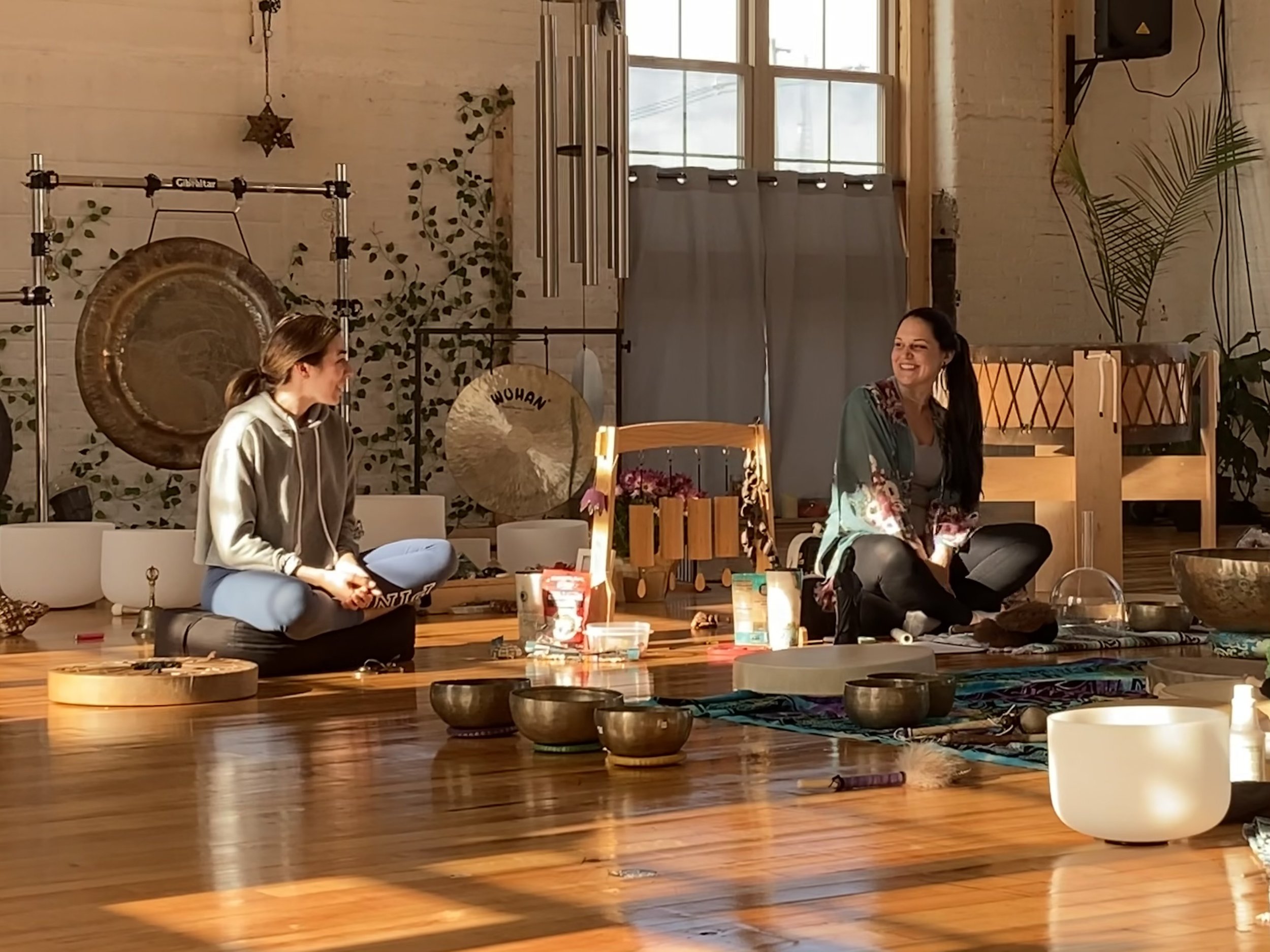 Two women sitting on the wooden floor of a meditation or sound therapy room, surrounded by singing bowls and musical instruments, smiling and engaging in conversation as sunlight streams through large windows.