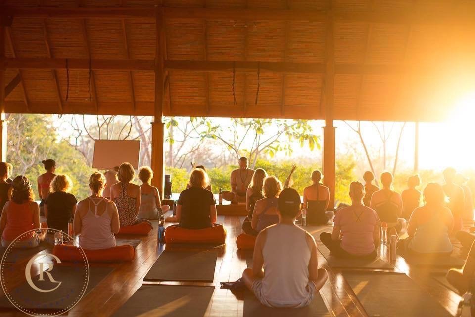 Group of people practicing yoga or meditation in a wooden, open-air space during sunrise or sunset.