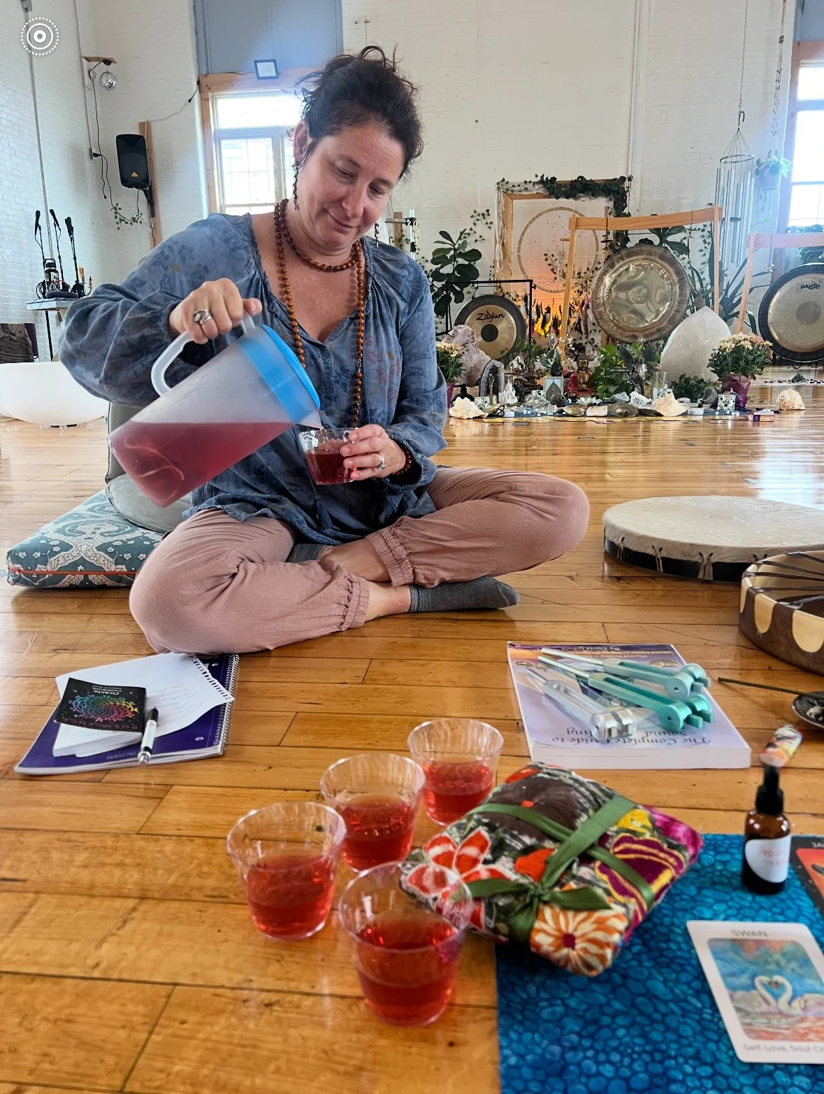 A woman sitting cross-legged on the floor pouring a red drink into a small glass, with four glasses of the drink in front of her. She is surrounded by notebooks, pens, a wrapped gift, and a small essential oil bottle. The setting appears indoors with musical instruments and spiritual items in the background.