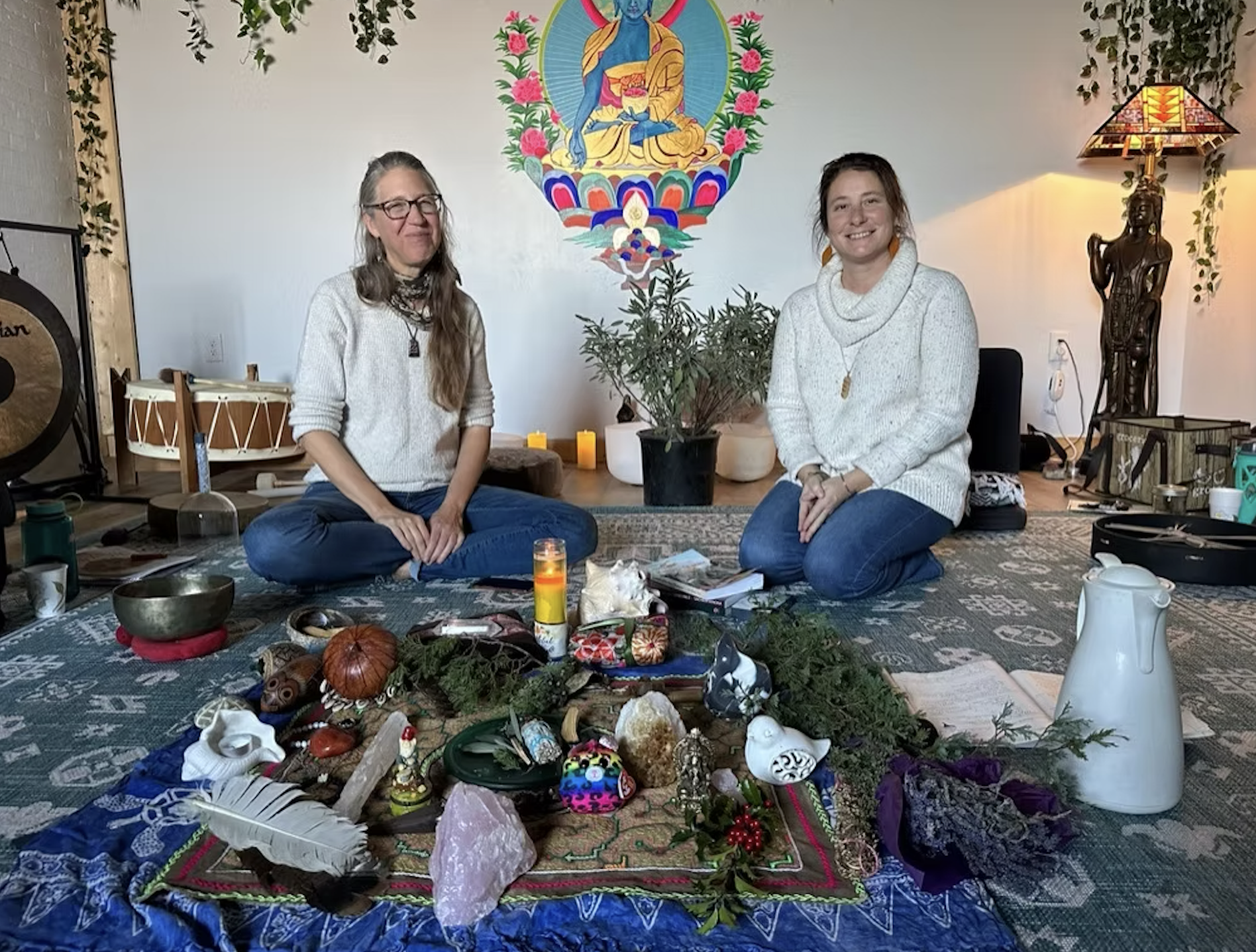 Two women sitting cross-legged on the floor in a yoga studio or spiritual space, surrounded by spiritual objects, with a colorful mat or cloth adorned with various items including crystals, candles, and small statues. Behind them, a large potted plant is visible, along with a vibrant, colorful painting of a seated figure on the wall and decor such as a lamp, statues, and candles.