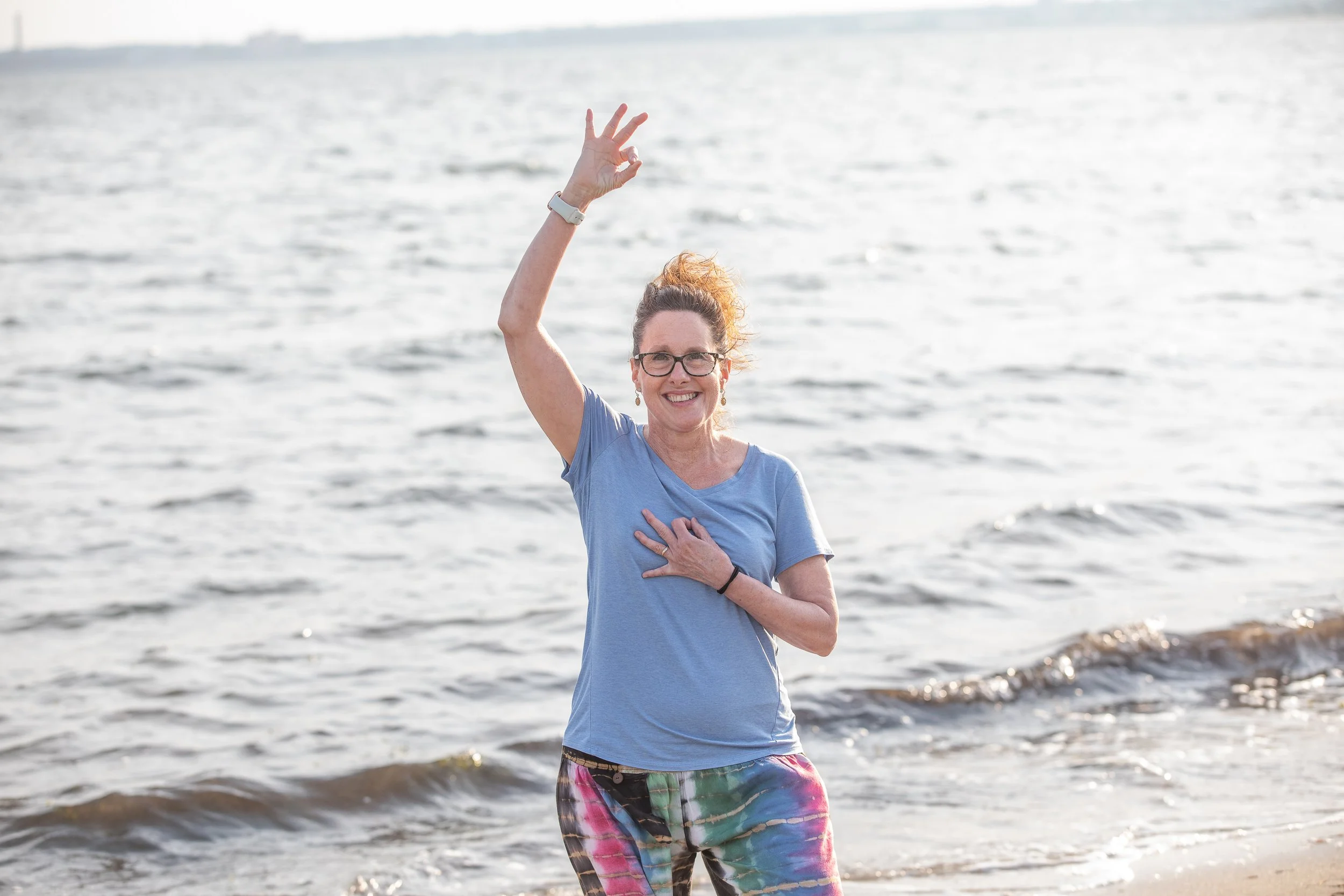 Woman standing at the beach raising her right hand, with her left hand on her chest, smiling, wearing glasses, a blue t-shirt, and colorful tie-dye pants.
