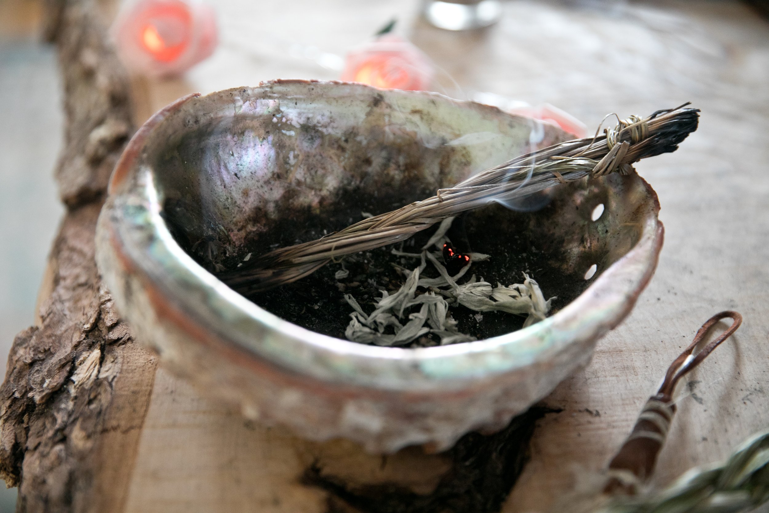 A ceramic bowl with burned herbs and incense stick on a wooden surface, with faint smoke rising. Sage burning to clear any density.