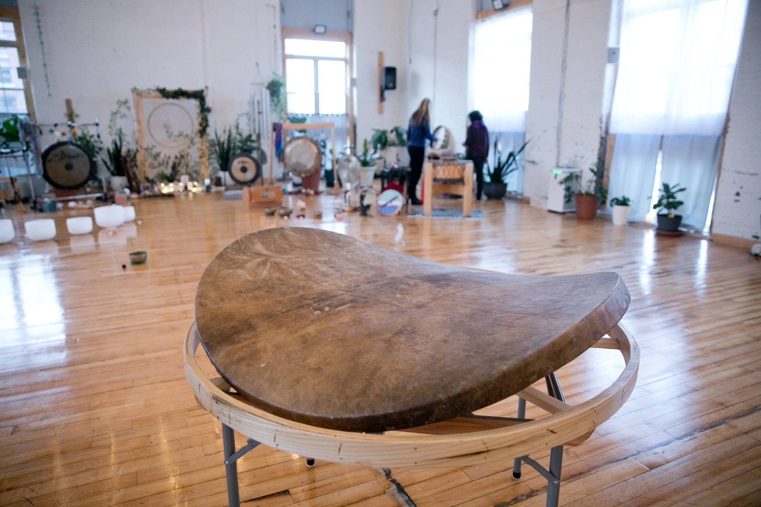A large wooden OHM Drum channeling device on a stand in a spacious room with wooden floors, potted plants, and people in the background of the yoga studio.
