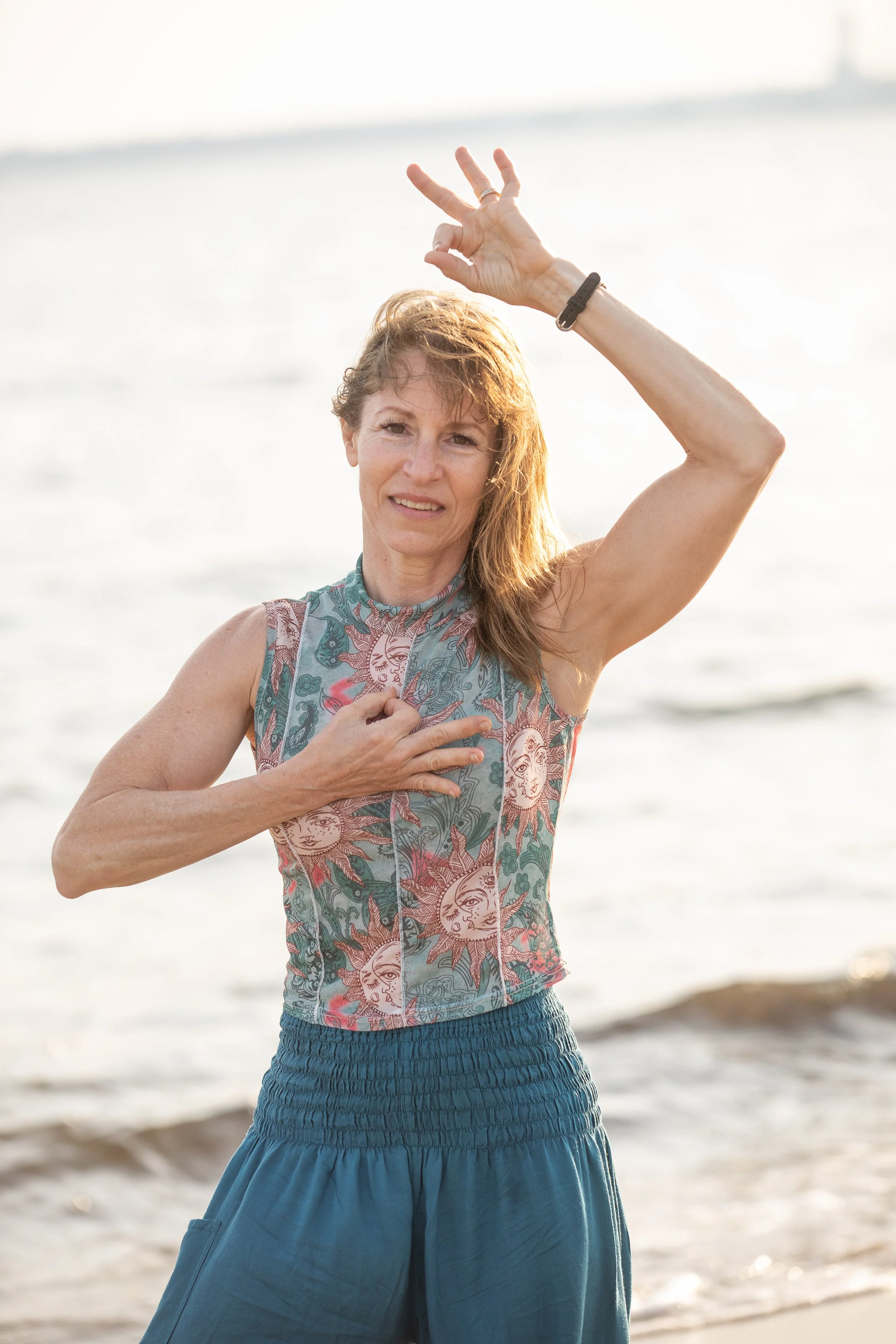 A woman standing on the beach during sunset, holding her hand to her chest and making a gesture with her other hand above her head.
