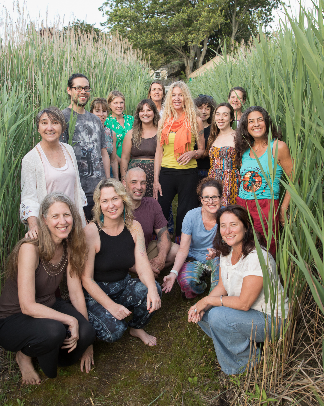 Group of people standing and sitting in a grassy outdoor area surrounded by tall green grass with trees in the background. Sva Ha Academy faculty beachside.
