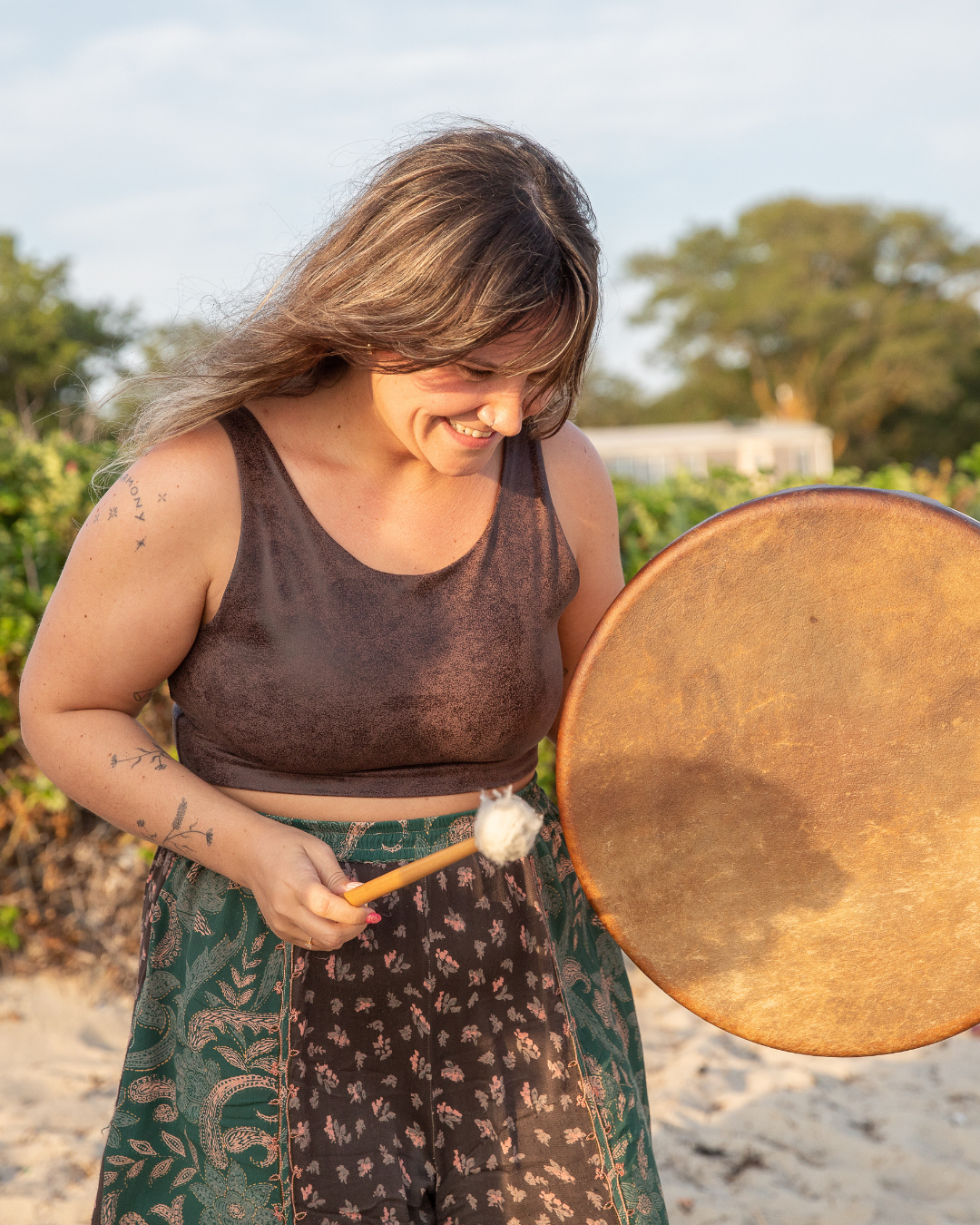 A woman smiling while holding a mallet and a large gong outdoors with greenery and trees in the background.