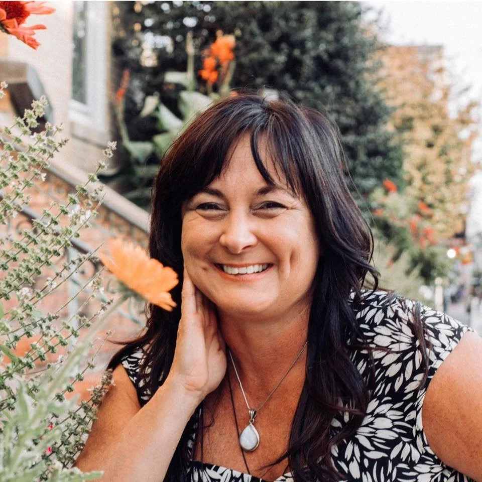 A woman with dark hair smiling outdoors among flowers and greenery.