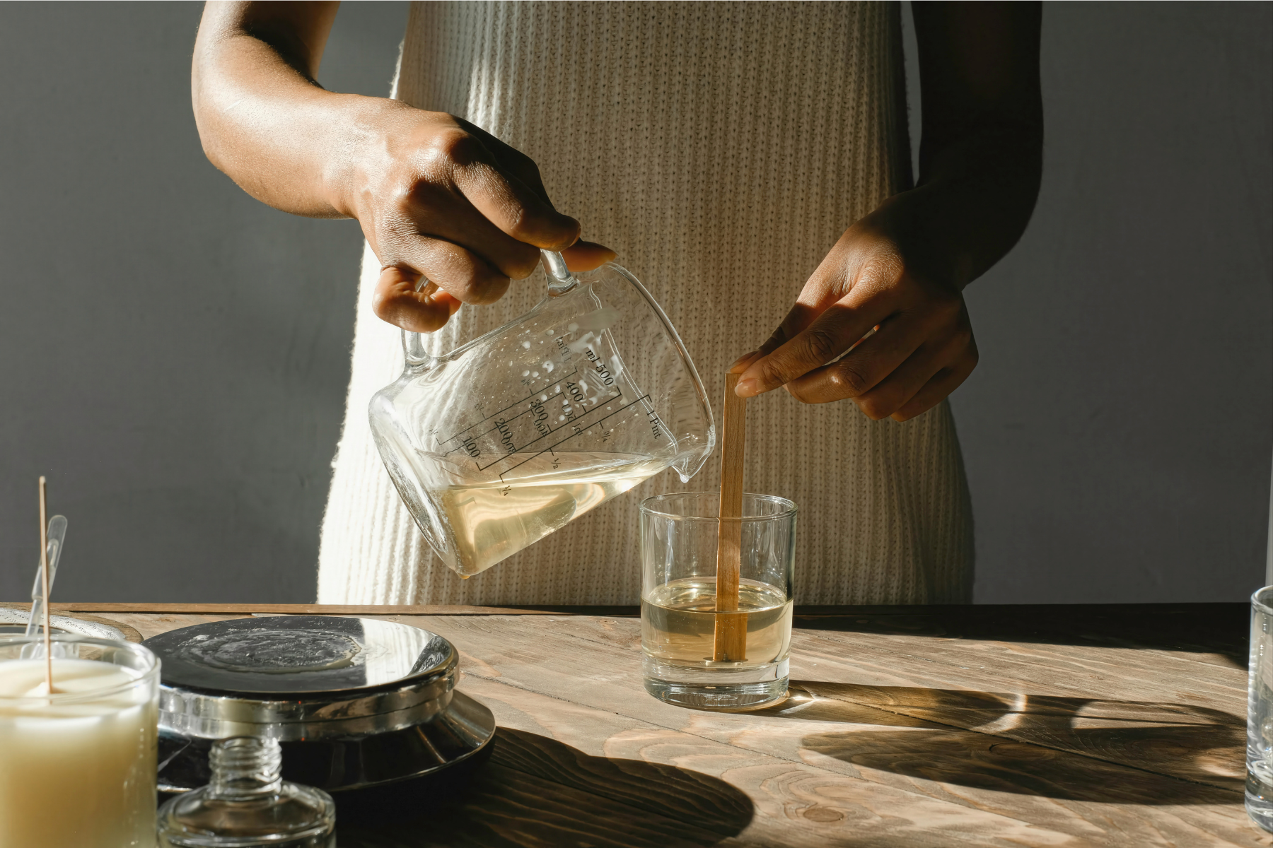 Person pouring liquid from a glass measuring cup into a glass container with a stirrer inside on a wooden table