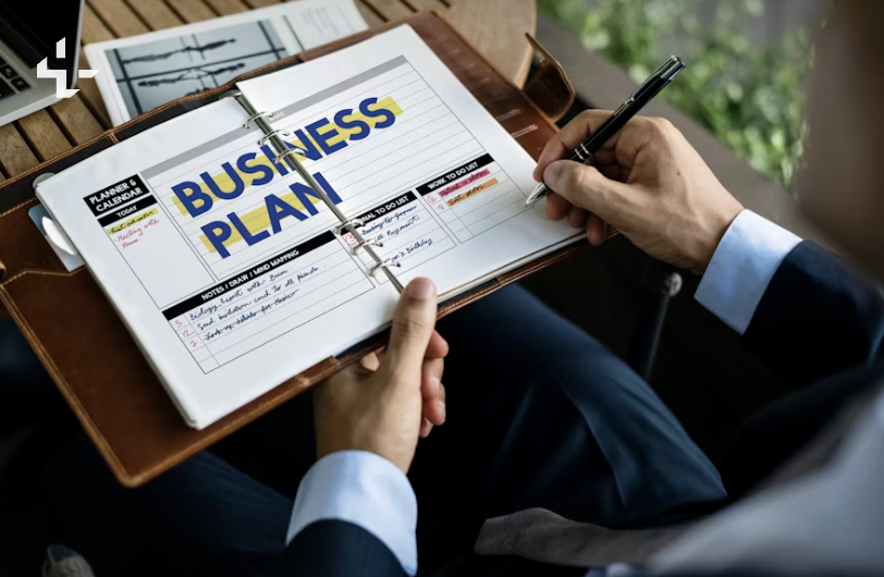Person in business attire writing in a planner with a business plan and notes, near a laptop and window with green plants.