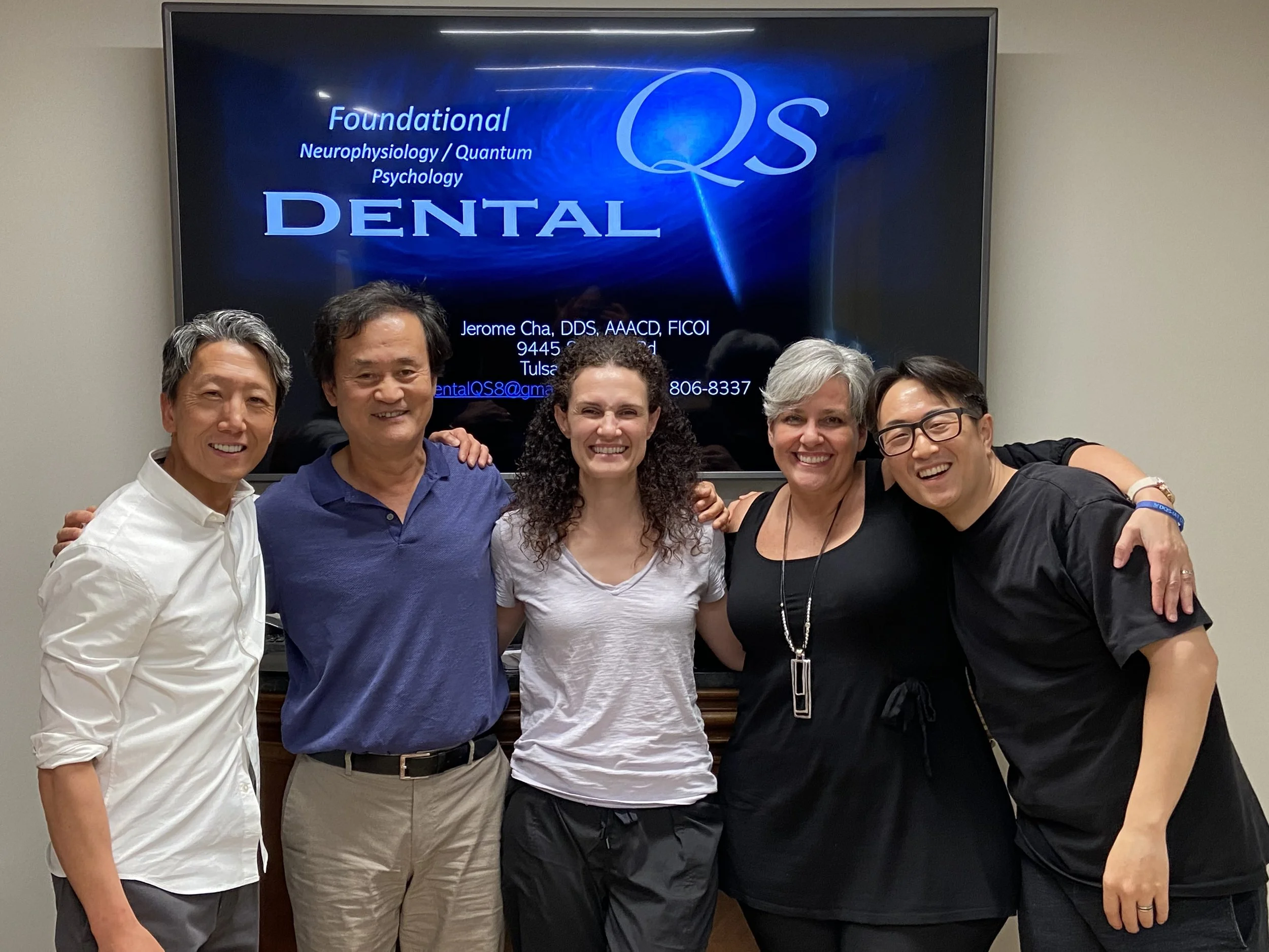 Group of five smiling people standing together in front of a presentation screen at a dental seminar, with a diverse mix of men and women, younger and older, in casual and semi-formal clothing.