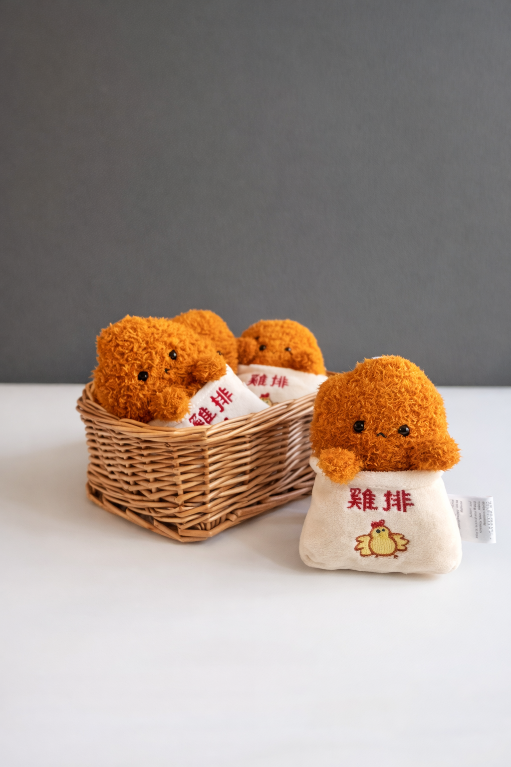 Four Ji Pai plush keychains in a brown woven basket — three stacked inside and one outside — in white embroidered “雞排” bags with cute eyes and 3-shaped smiles.