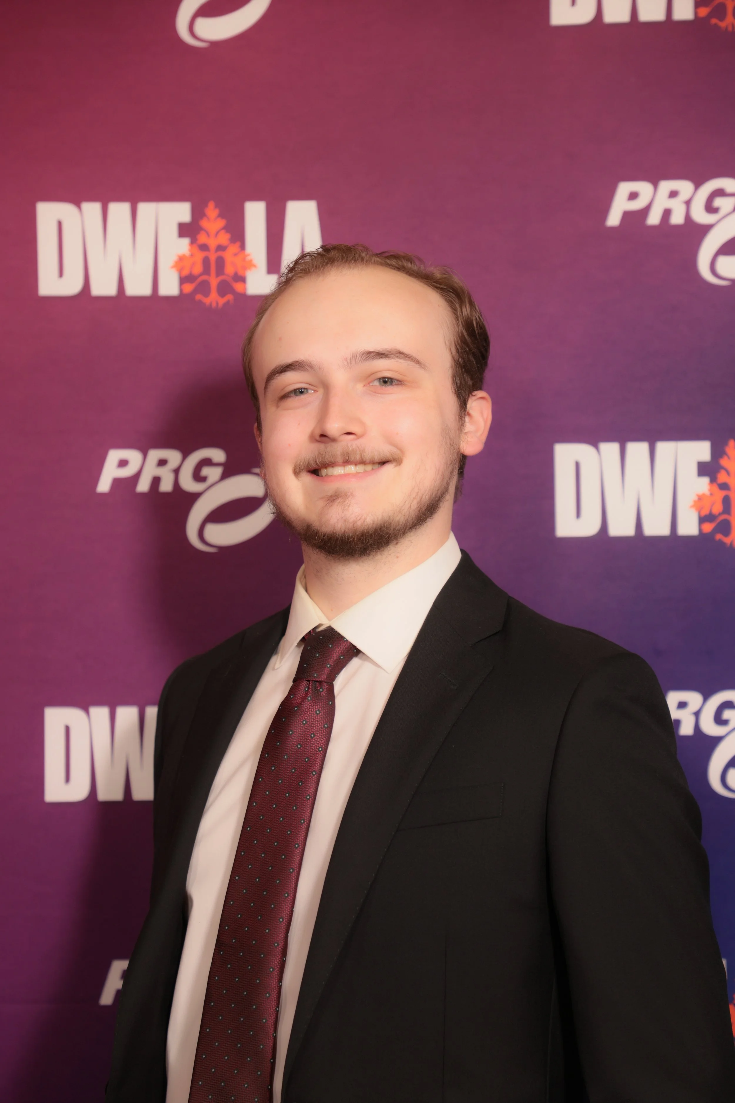 Young man in a black suit and red polka dot tie smiling, standing in front of a purple backdrop with logos and text.