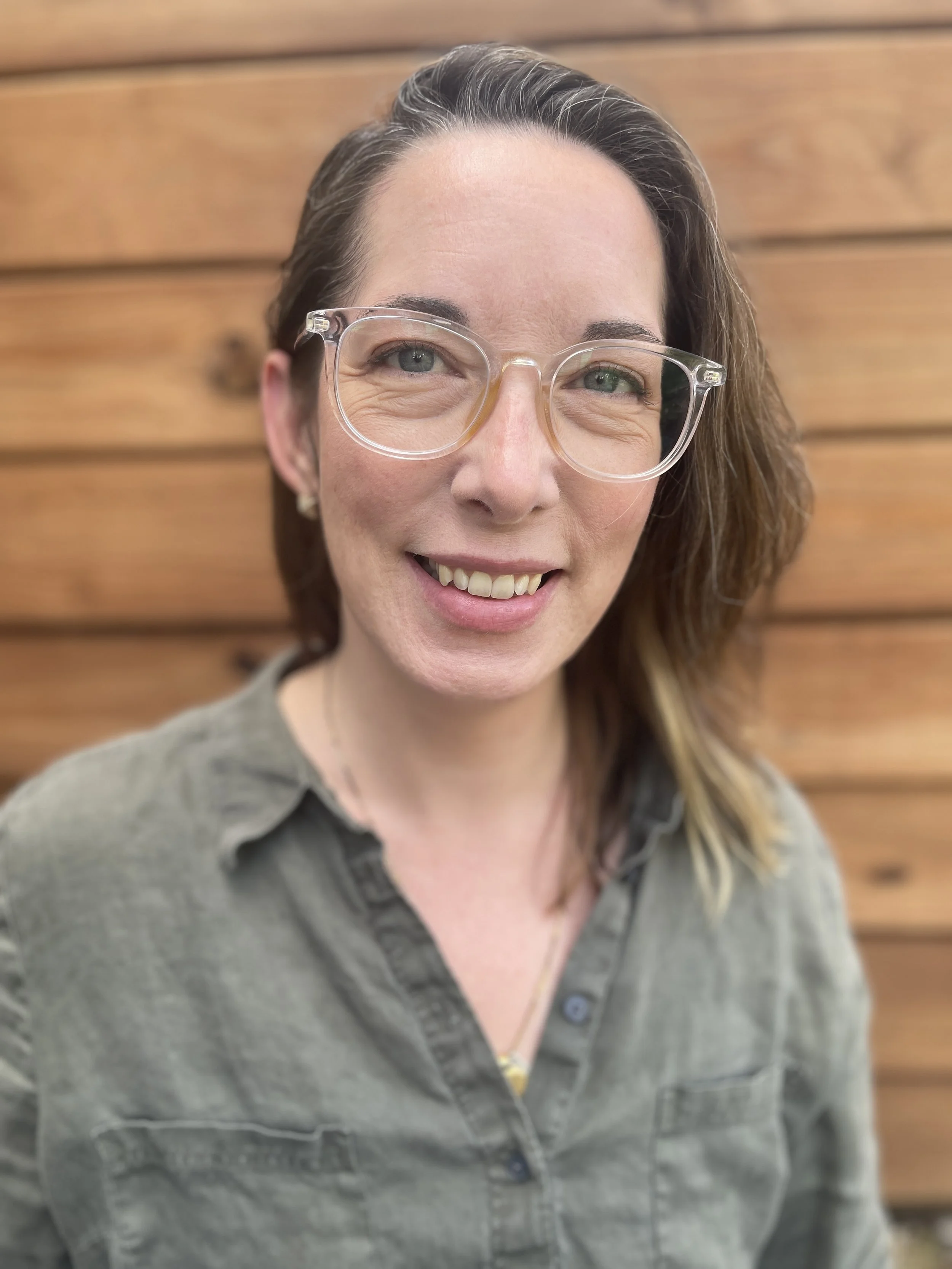 Close-up of a woman with light skin, glasses, and long wavy brown hair, smiling, against a wooden background.