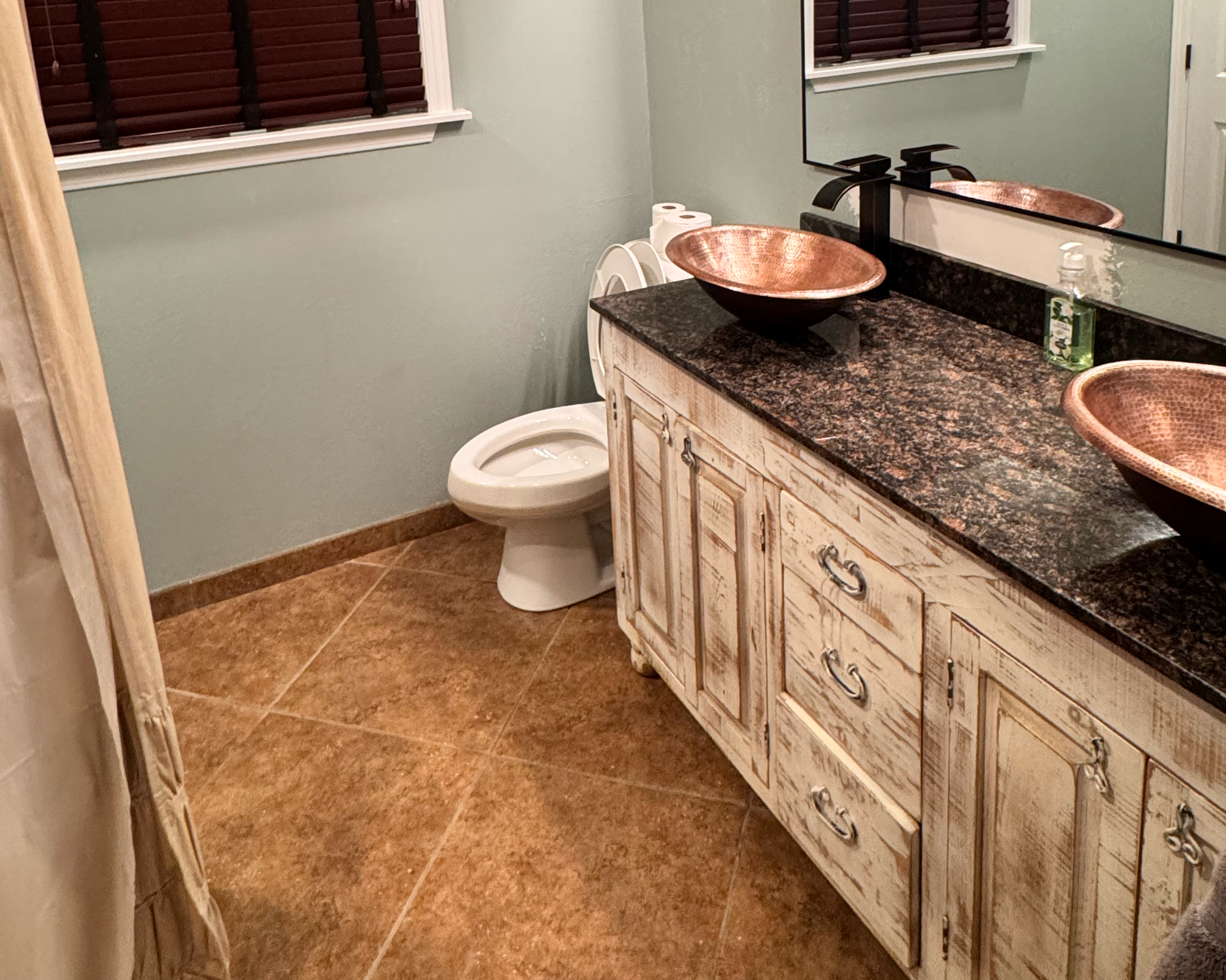 Bathroom with green walls, dark wooden blinds on window, mirror, light fixture, vanity with granite countertop, copper bowls, small decorative items, and a toilet.