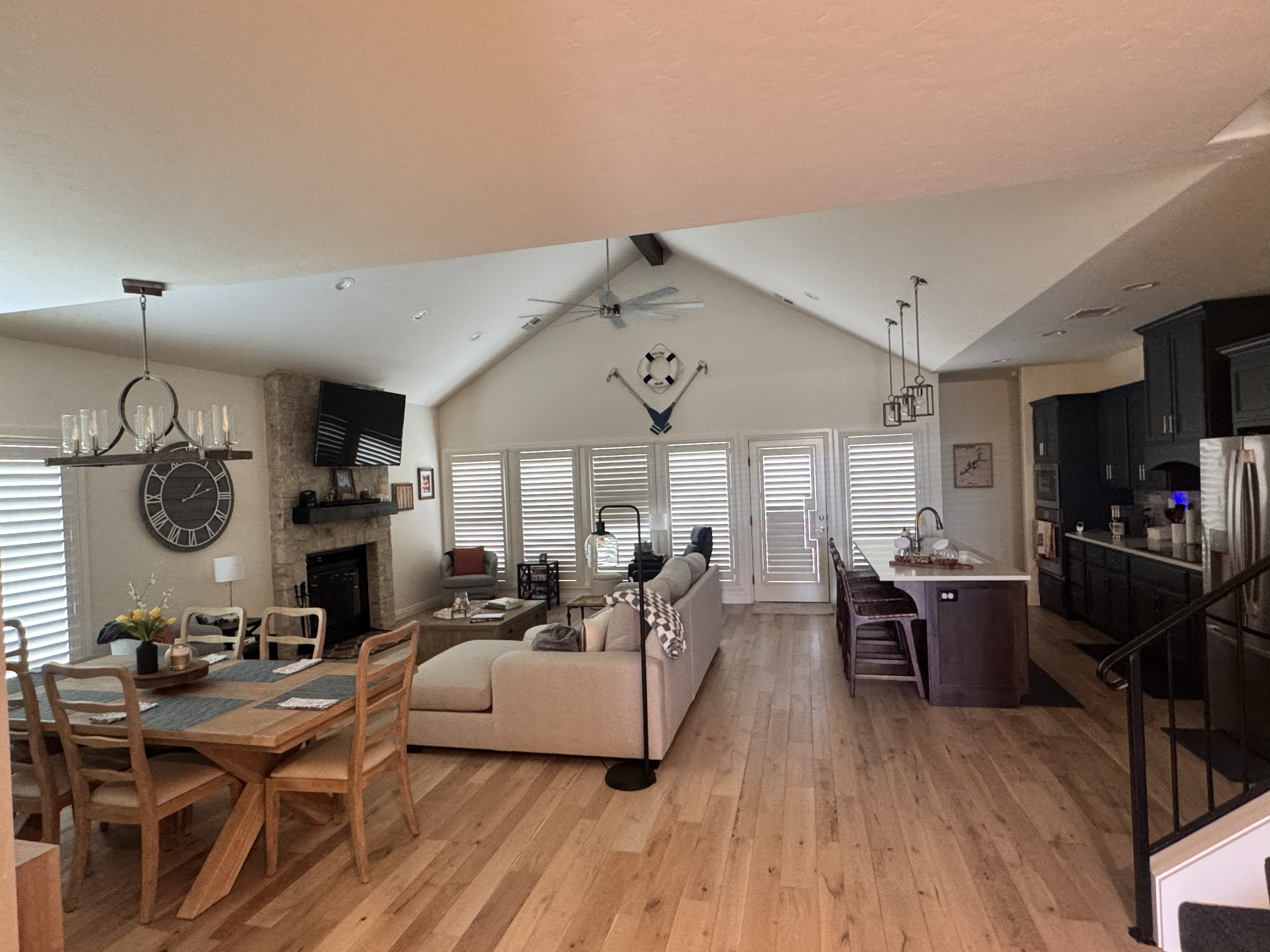 Open-concept living room and kitchen with hardwood floors, white walls, ceiling fan, clock, fireplace, flat screen TV, dining area, and kitchen island with bar stools.
