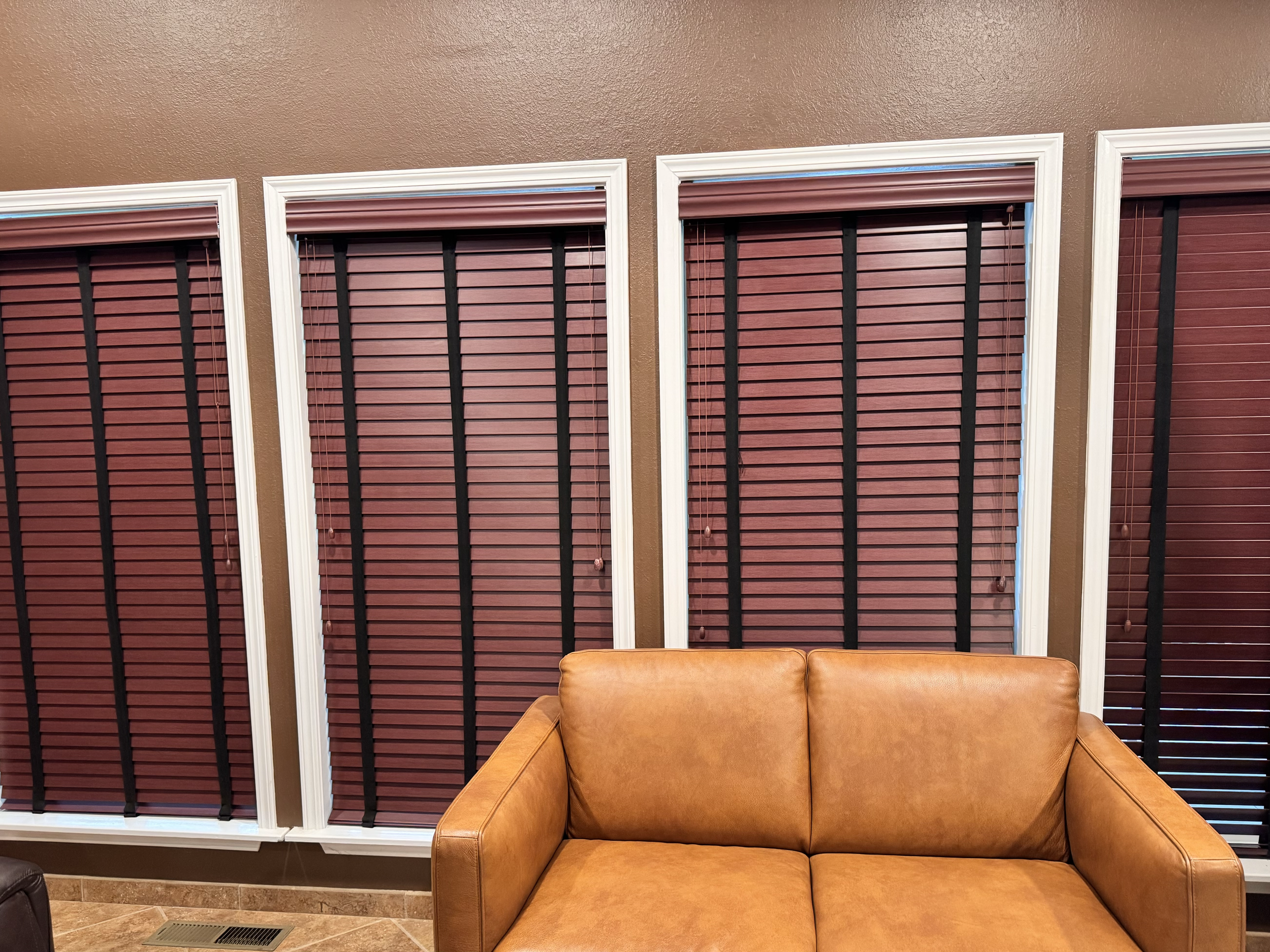 Living room with three windows covered with pinkish-red blinds and a tan leather sofa in the foreground.