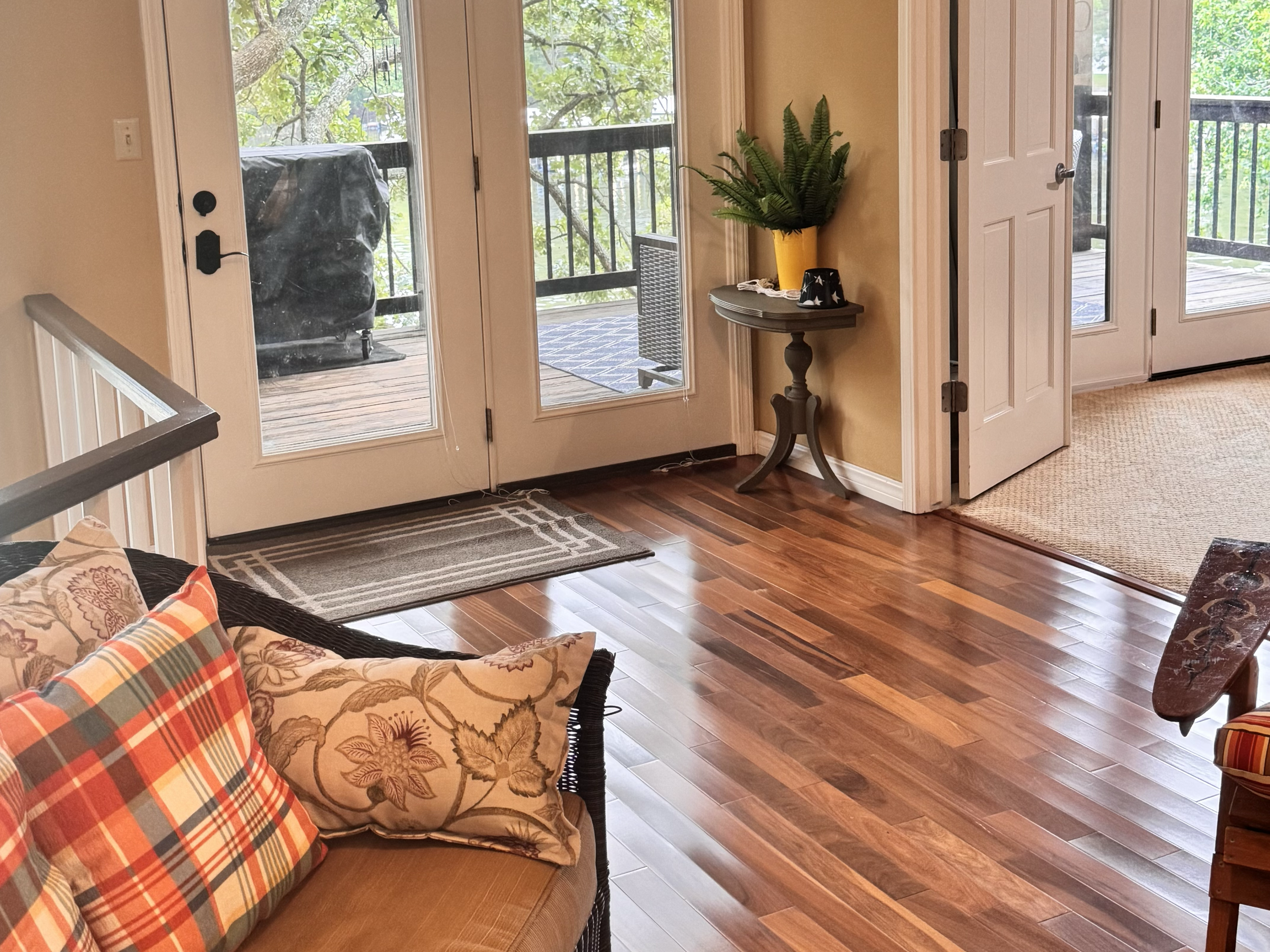 Living room with wood flooring, a black wicker sofa with plaid and floral pillows, glass double doors leading to a balcony with trees visible outside, a small round side table holding a plant in a yellow pot and two cups, and a beige carpeted area ou