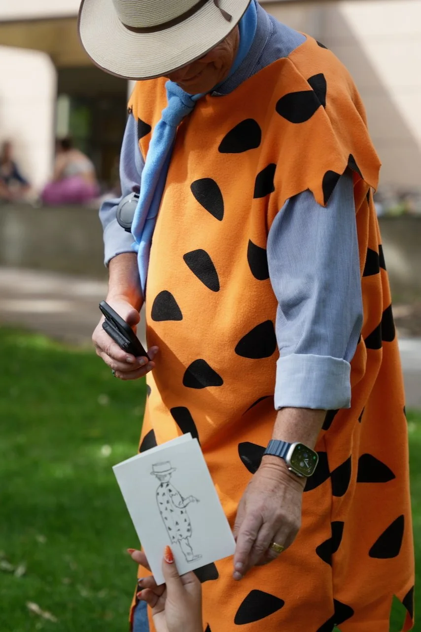 A person dressed as Fred Flintstone from The Flintstones is standing outside, wearing an orange caveman costume with black spots, a blue long-sleeved shirt, and a white hat with a black band. Someone else is handing the person a small card with a sketch of Fred Flintstone on it.