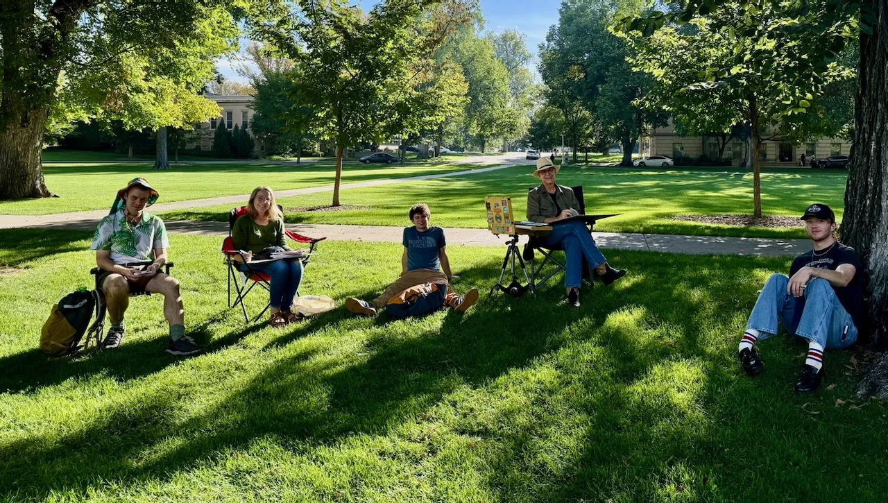 A group of five people during a painting session sitting on the grass in a park on a sunny day, with trees and a building in the background.