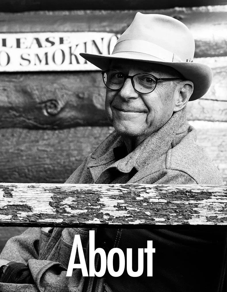 A black and white photo of a smiling man wearing glasses and a fedora hat, seated outdoors with a wooden background and a sign that reads 'Please No Smoking' in the background.