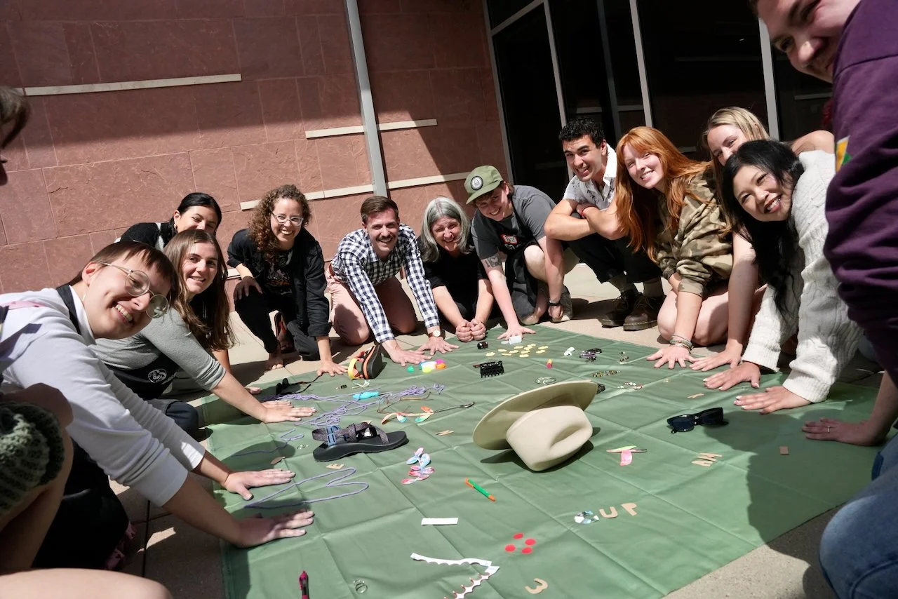 Group of diverse people smiling and gathered around a green cloth with various trinkets and objects, inside a building with red brick walls.