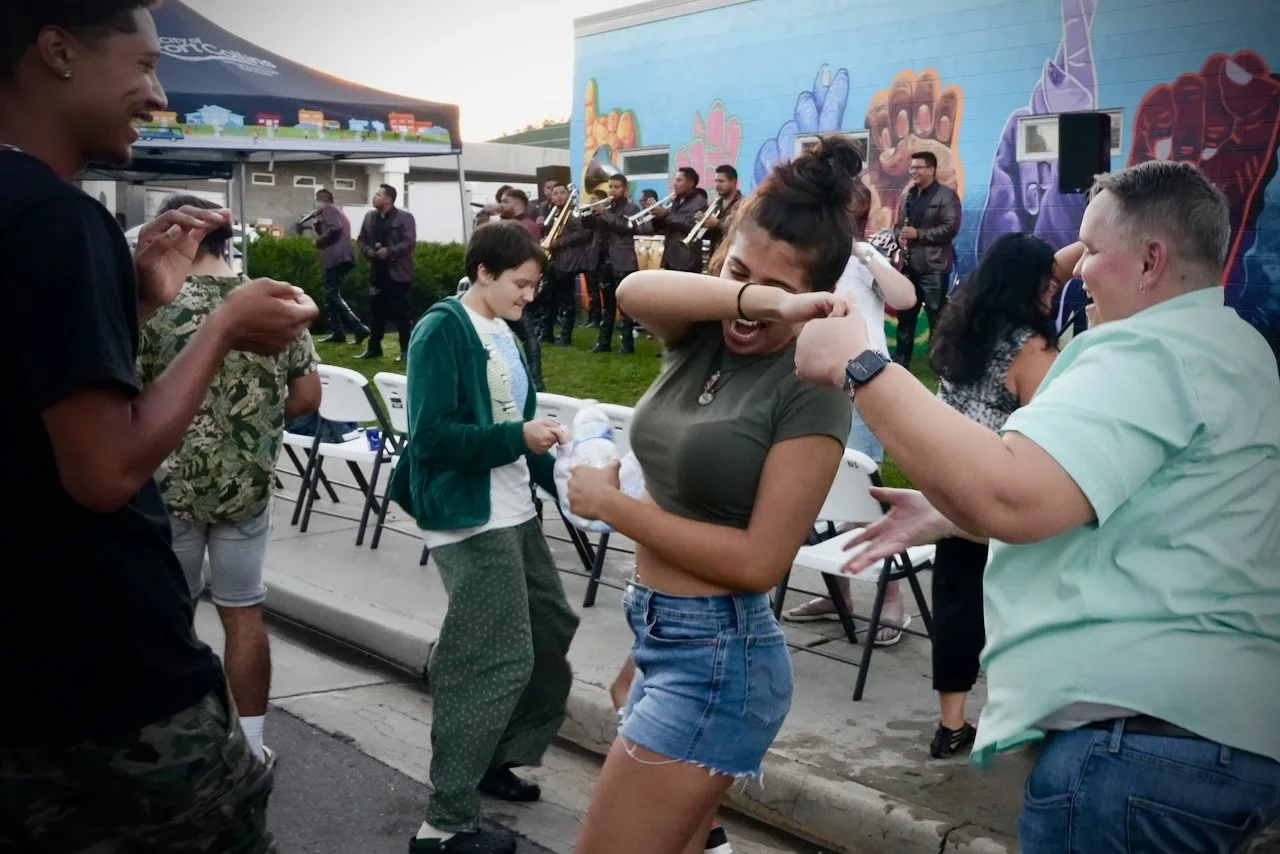 Group of people dancing and enjoying music outdoors at a lively event, with a band playing in the background and colorful wall art.