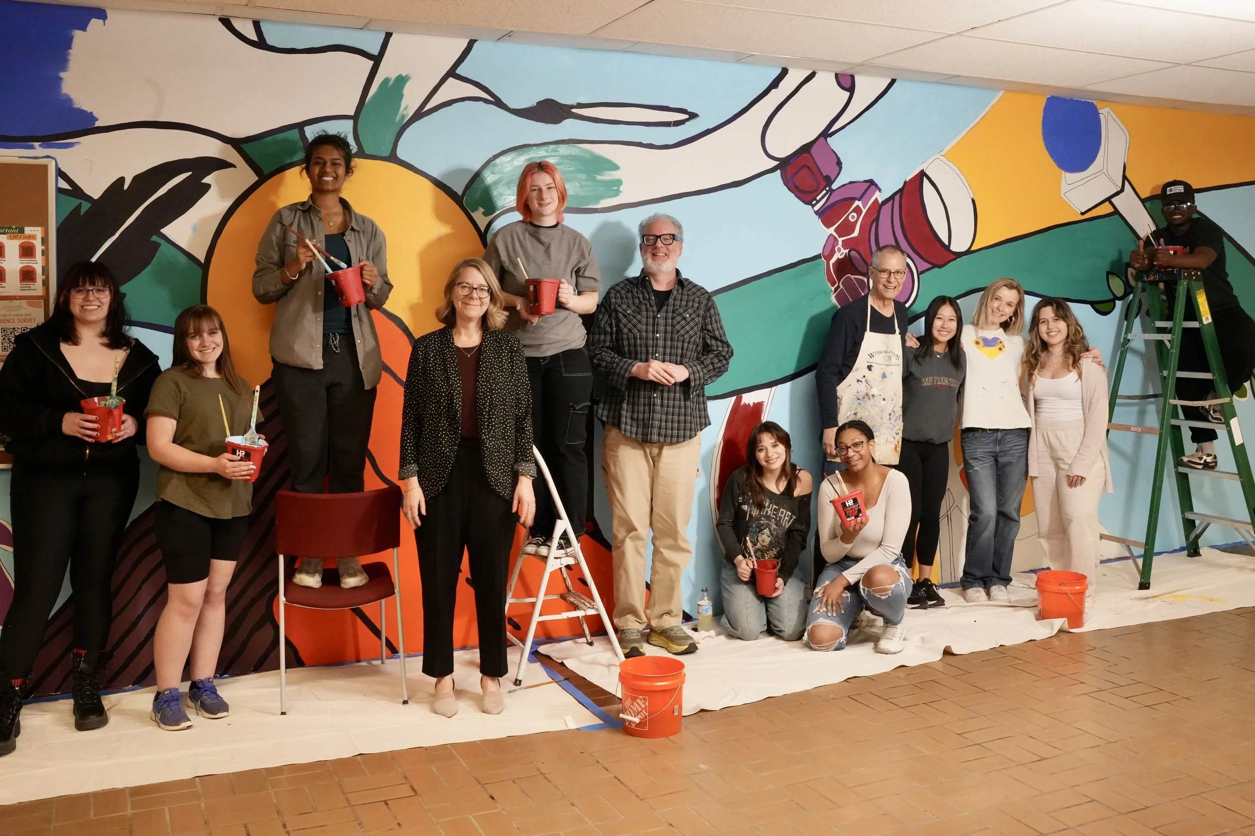 Group of people painting a colorful mural on a wall, some holding paint buckets and brushes, with ladders and paint supplies around them.