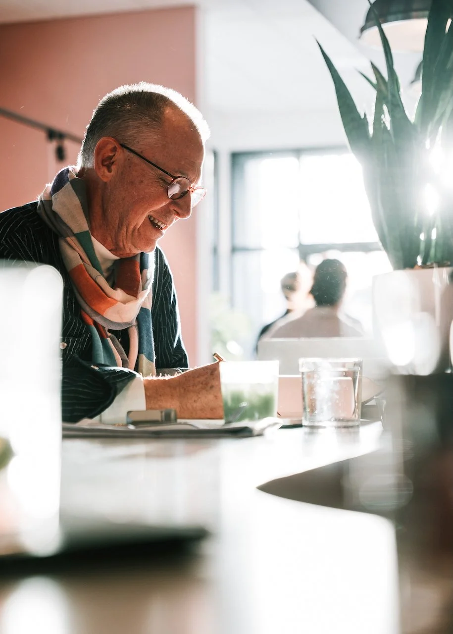 David Pyle smiling and writing at a table in a bright, modern cafe, with plants and sunlight streaming through large windows.