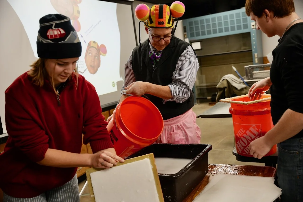 Three people working with soap making supplies, one woman pouring soap mixture from a bucket into a mold, a man wearing a colorful mouse hat, and another woman holding a tray and measuring hot liquid.