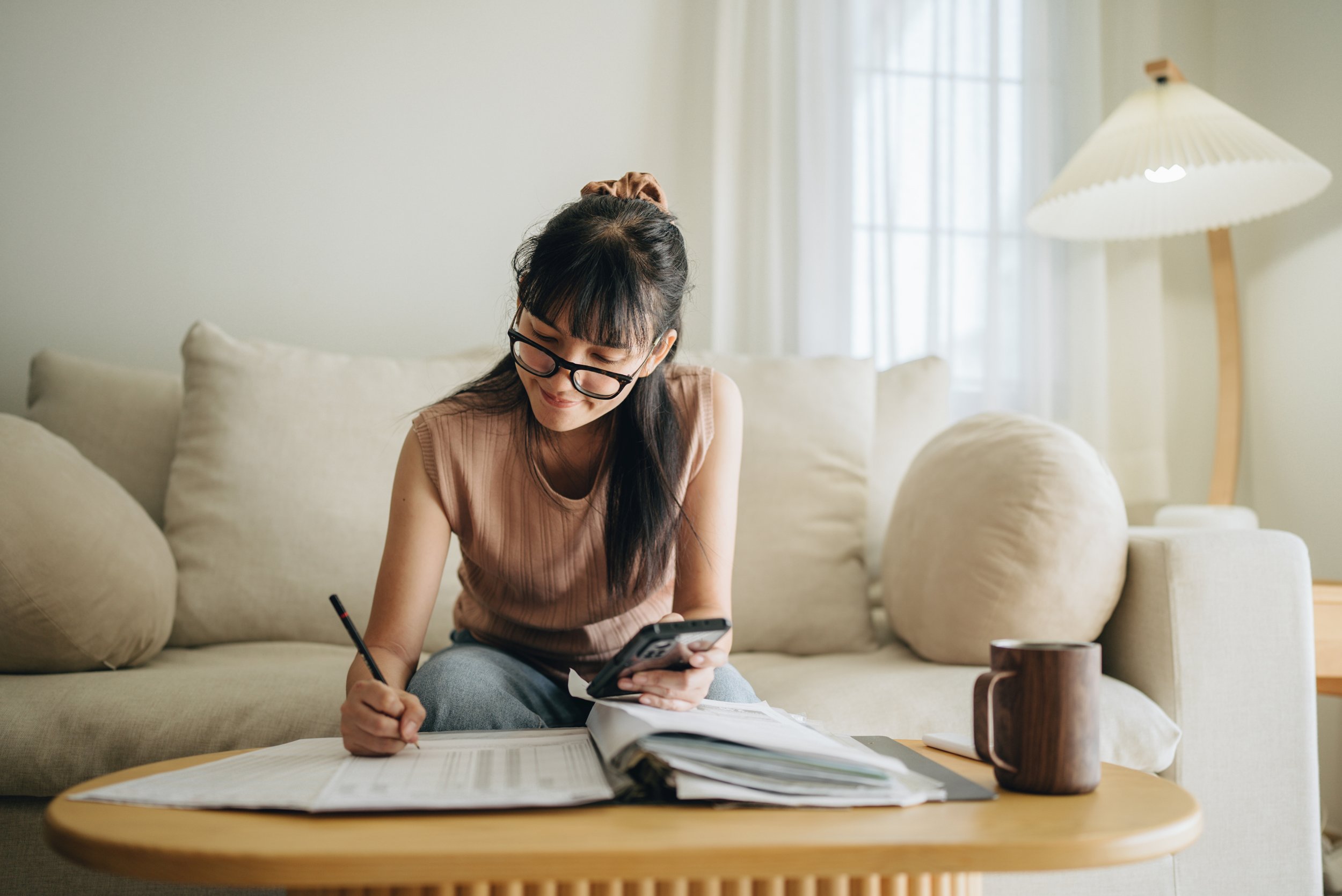 Woman sits on couch, looking at papers and writing.