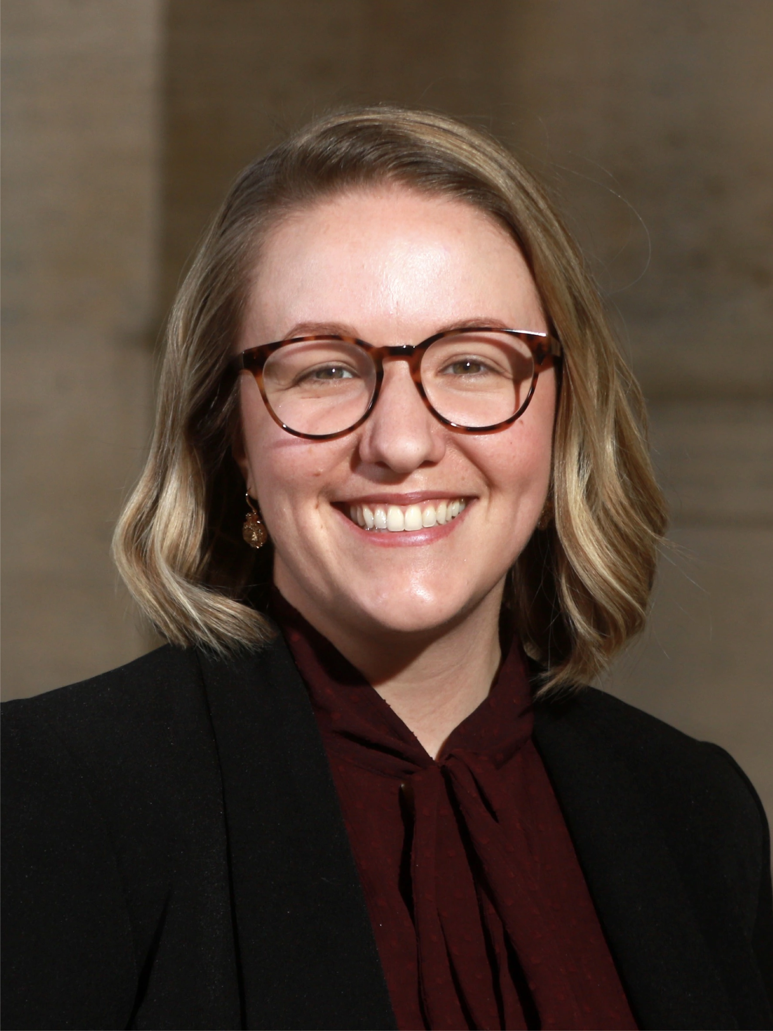 A smiling woman with shoulder-length blonde hair, glasses, black blazer, and maroon blouse against a blurred background.